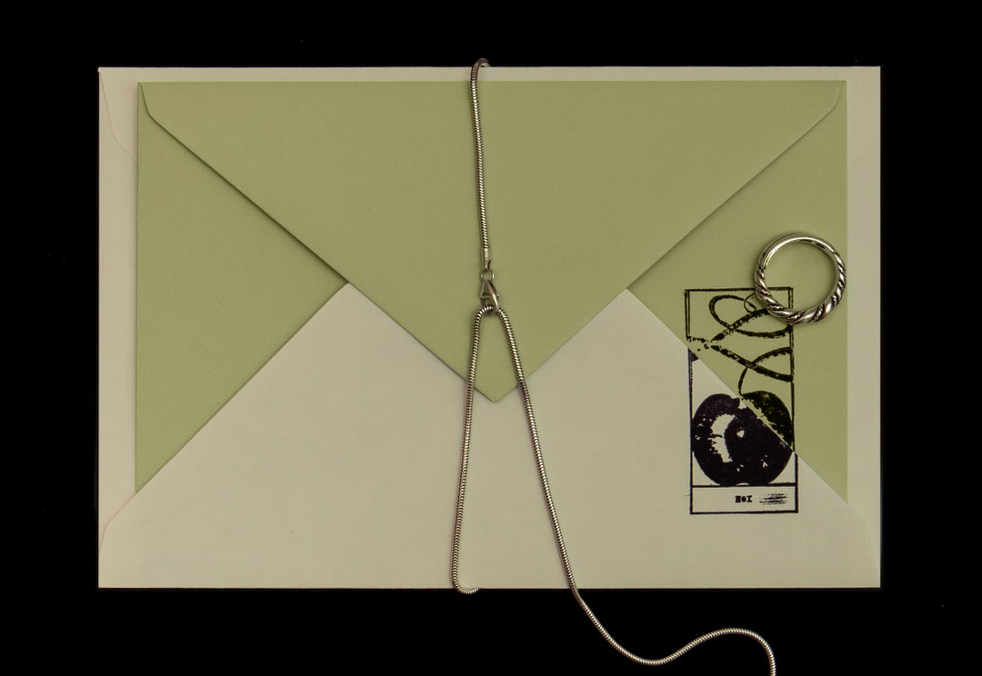 A pale green envelope rests on a cream background, its flap open. A silver snake chain is draped across the centre, paired with a twisted silver ring and a small black and white illustration of a fruit.