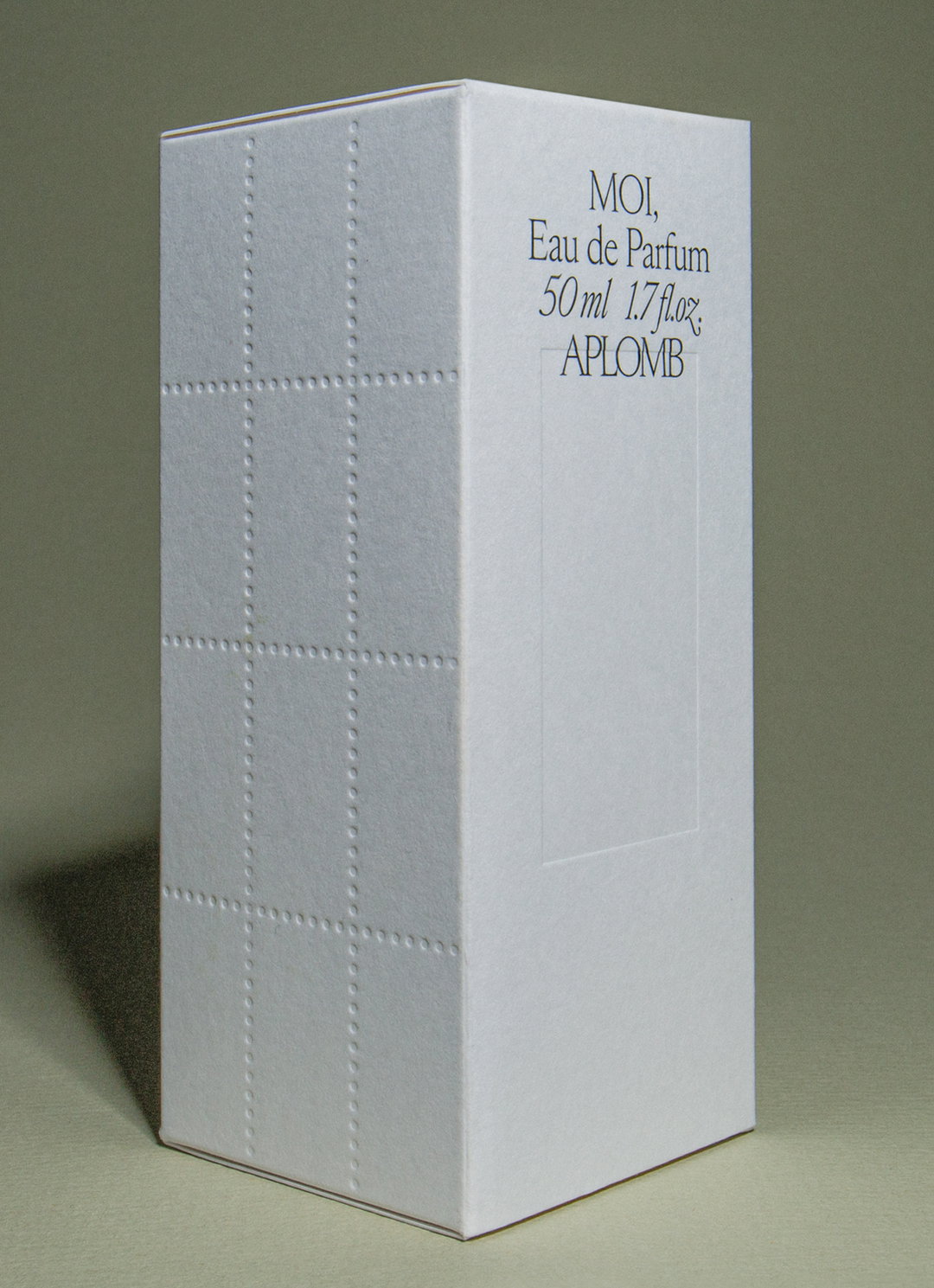 A tall, white rectangular perfume box stands against a neutral grey background. Text on the front reads “MOI, Eau de Parfum 50 ml 1.7 fl.oz, APLOMB”.