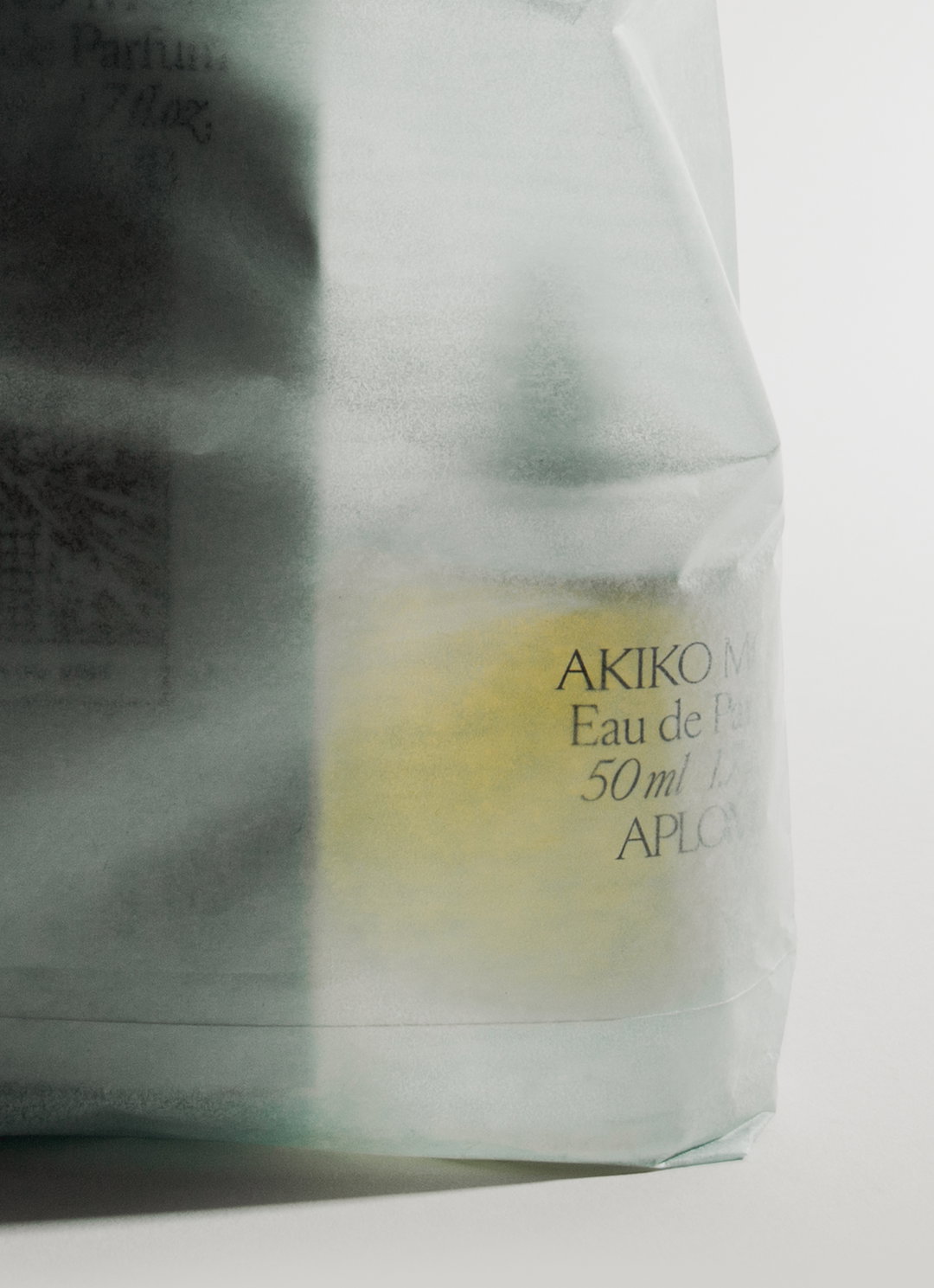 A translucent, frosted glass bottle contains a pale yellow liquid. Text on the side reads “AKIKO M”, “Eau de Par”, “50 ml” and “APLON”.