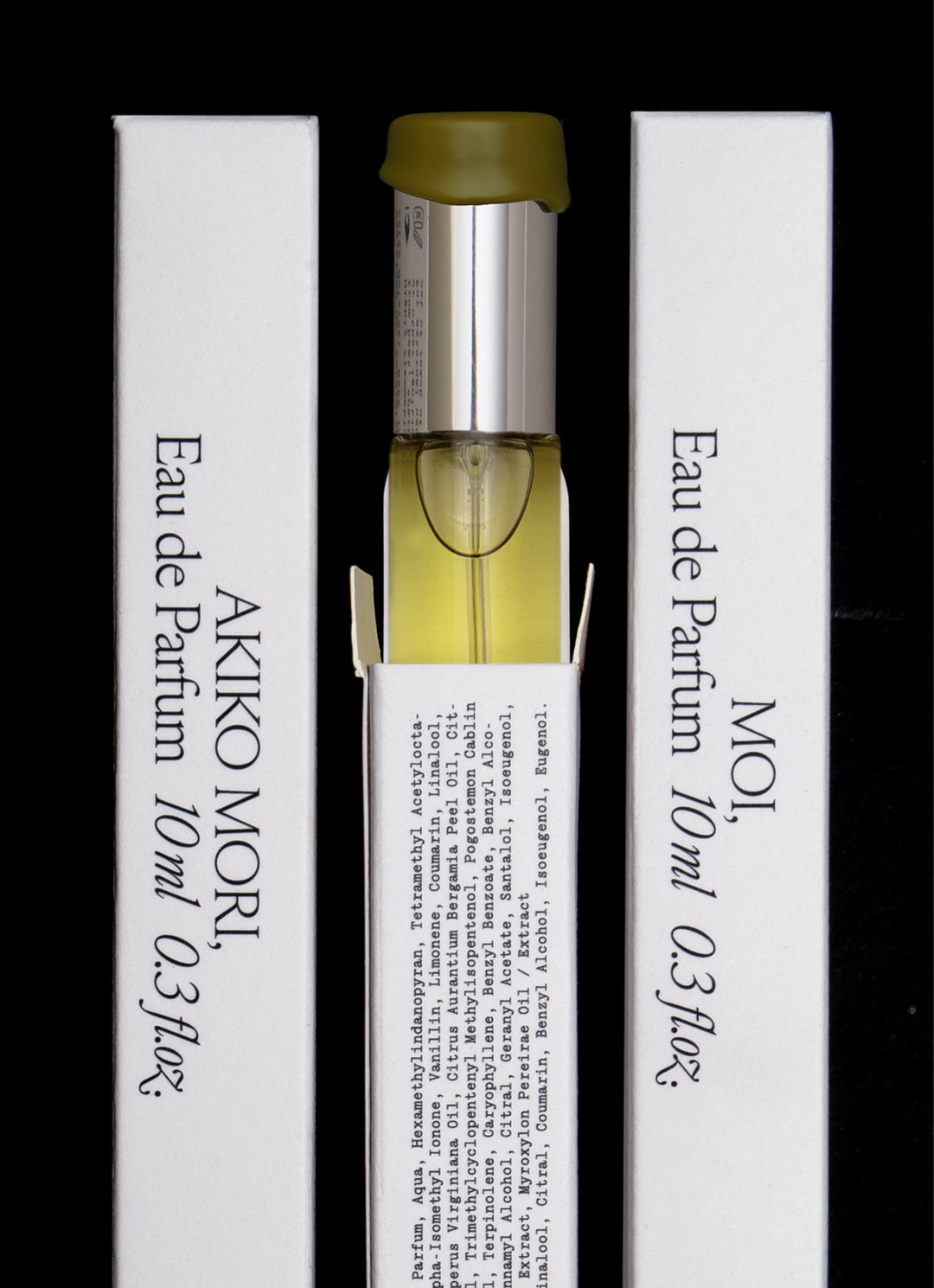 A small bottle of yellow perfume sits between two white cardboard packaging sleeves. The sleeves feature black text that reads “AKIKO MORI, Eau de Parfum 10ml 0.3fl.oz” and “MOI, Eau de Parfum 10ml 0.3fl.oz”.