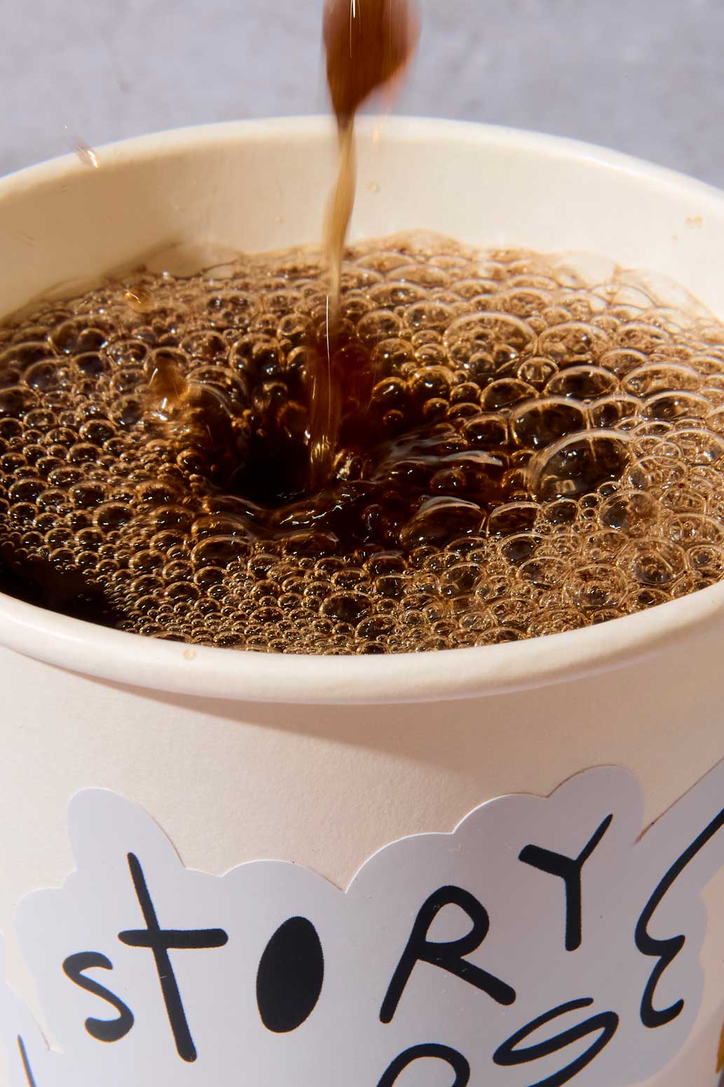 Dark coffee pours into a white paper cup, creating a layer of small brown bubbles on the surface. A white sticker on the cup features the word “story” in hand-drawn black lettering.