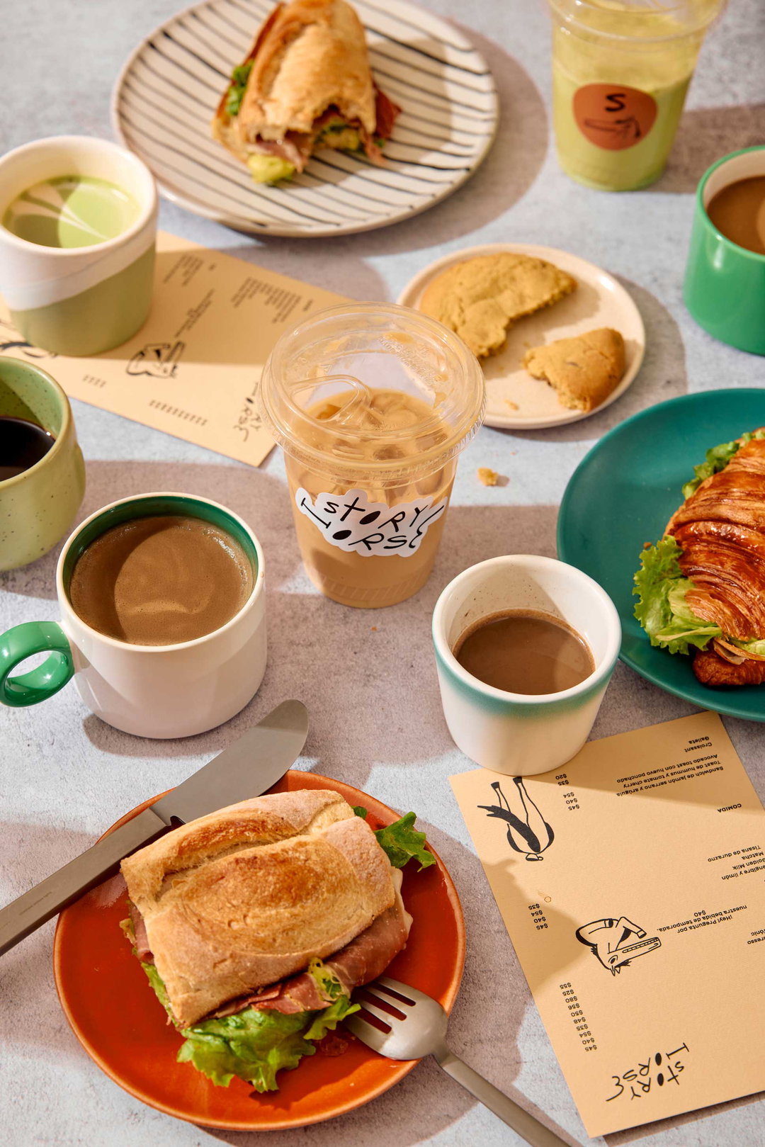 A spread of breakfast food and drinks on a grey table. The scene includes two croissants filled with lettuce and meat, a broken cookie, and several coffees and matcha lattes. One plastic cup features a white sticker that reads “Story Roast”.