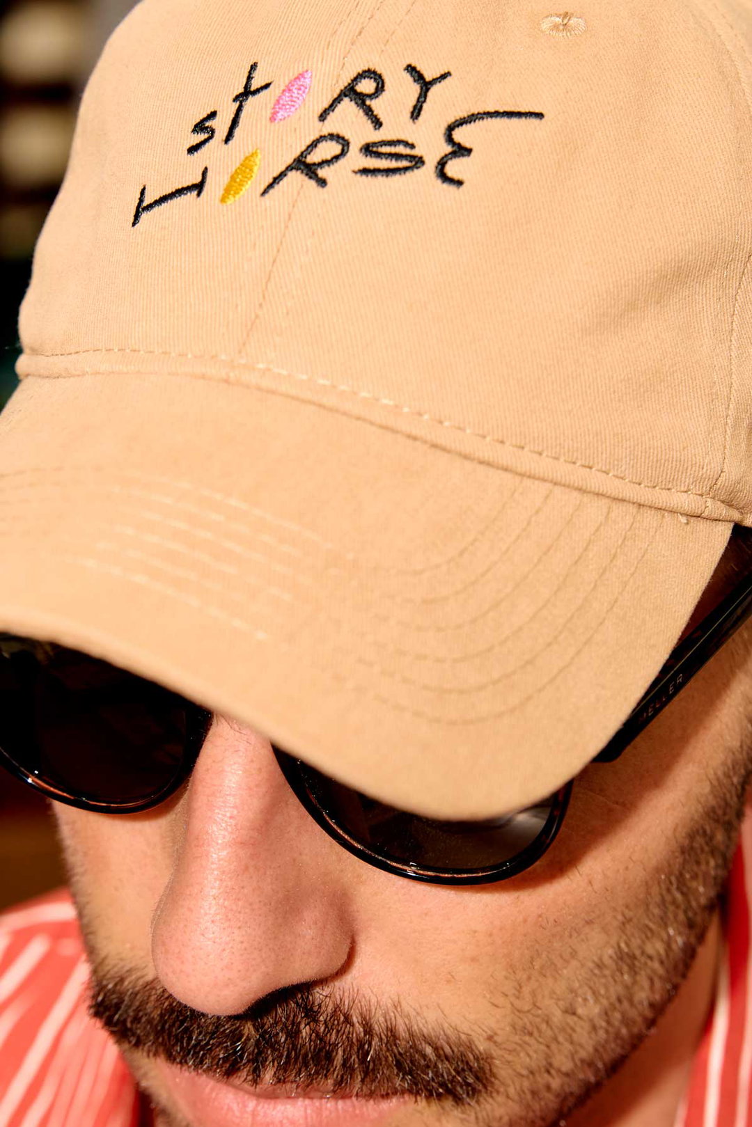 A close-up of a person wearing a beige baseball cap and dark sunglasses. The cap features the words “story horse” embroidered in a playful, multi-coloured font.