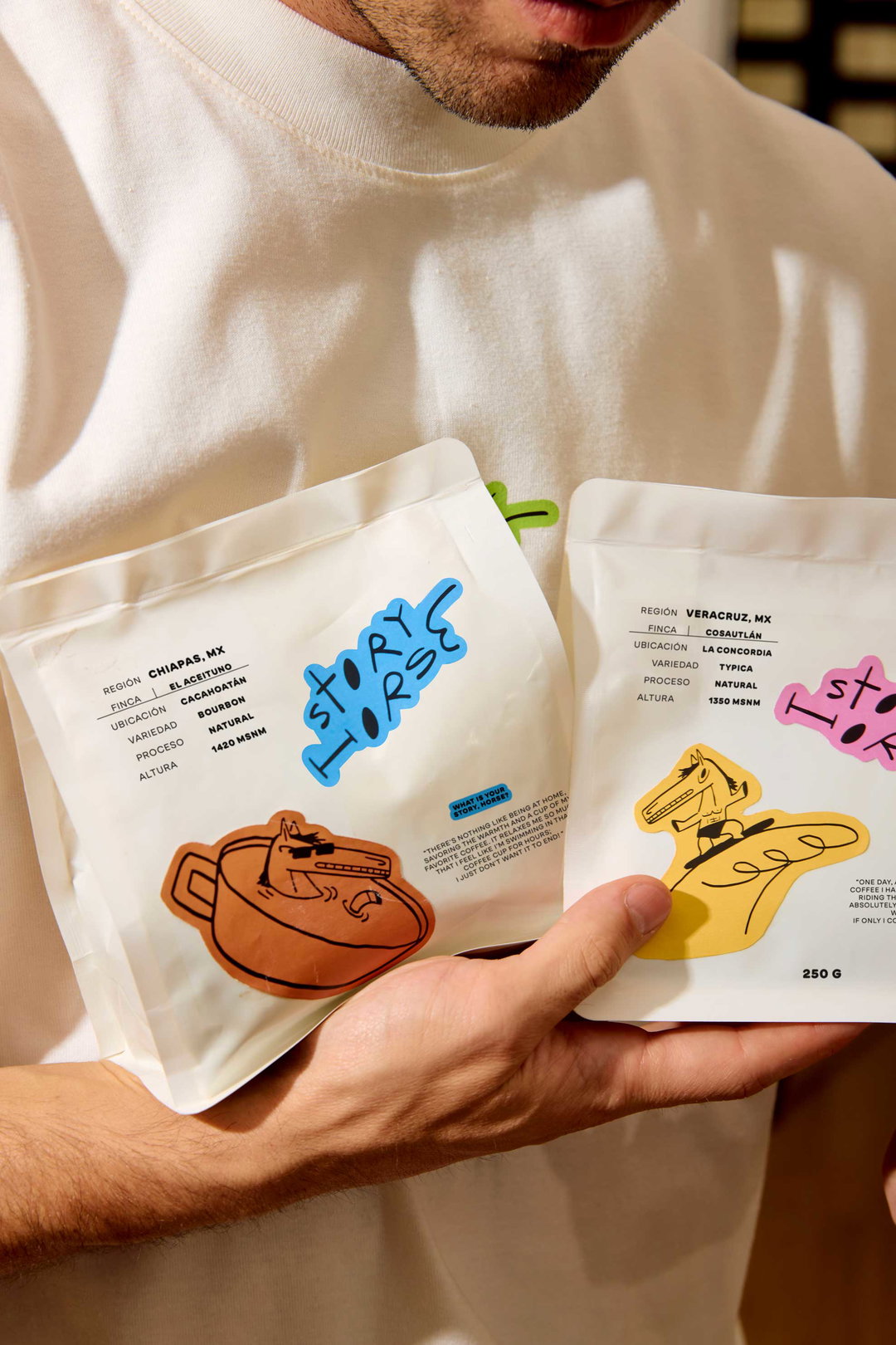 A person holds two white coffee bags with colourful, playful branding. One bag features a blue “Story Horse” logo and an illustration of a brown pot, while the other shows a pink logo and a yellow sketch of a horse.