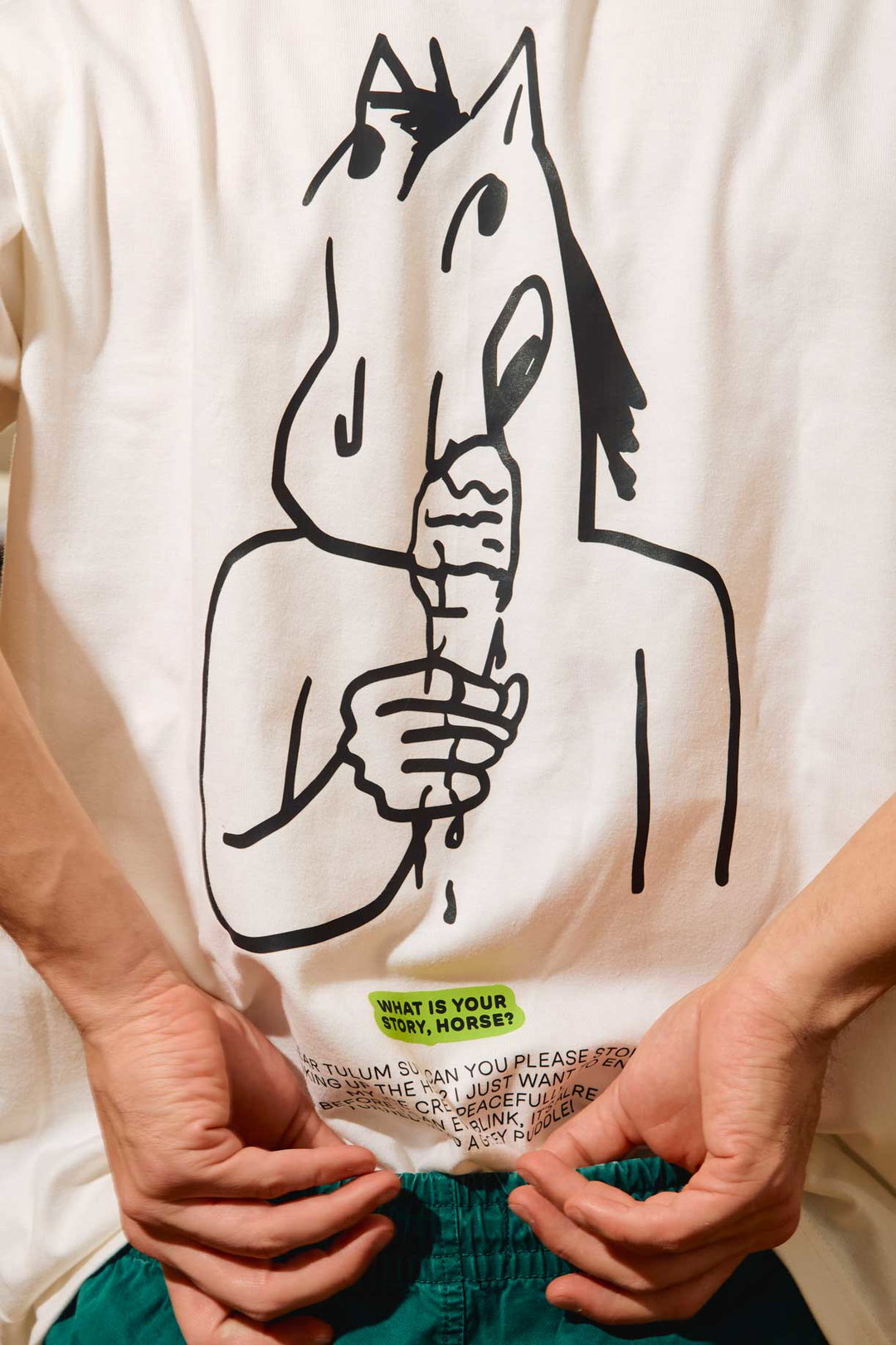 A person wears a white T-shirt featuring a black line drawing of a figure with a horse's head eating an ice cream. A small green label at the bottom reads “WHAT IS YOUR STORY, HORSE?”.