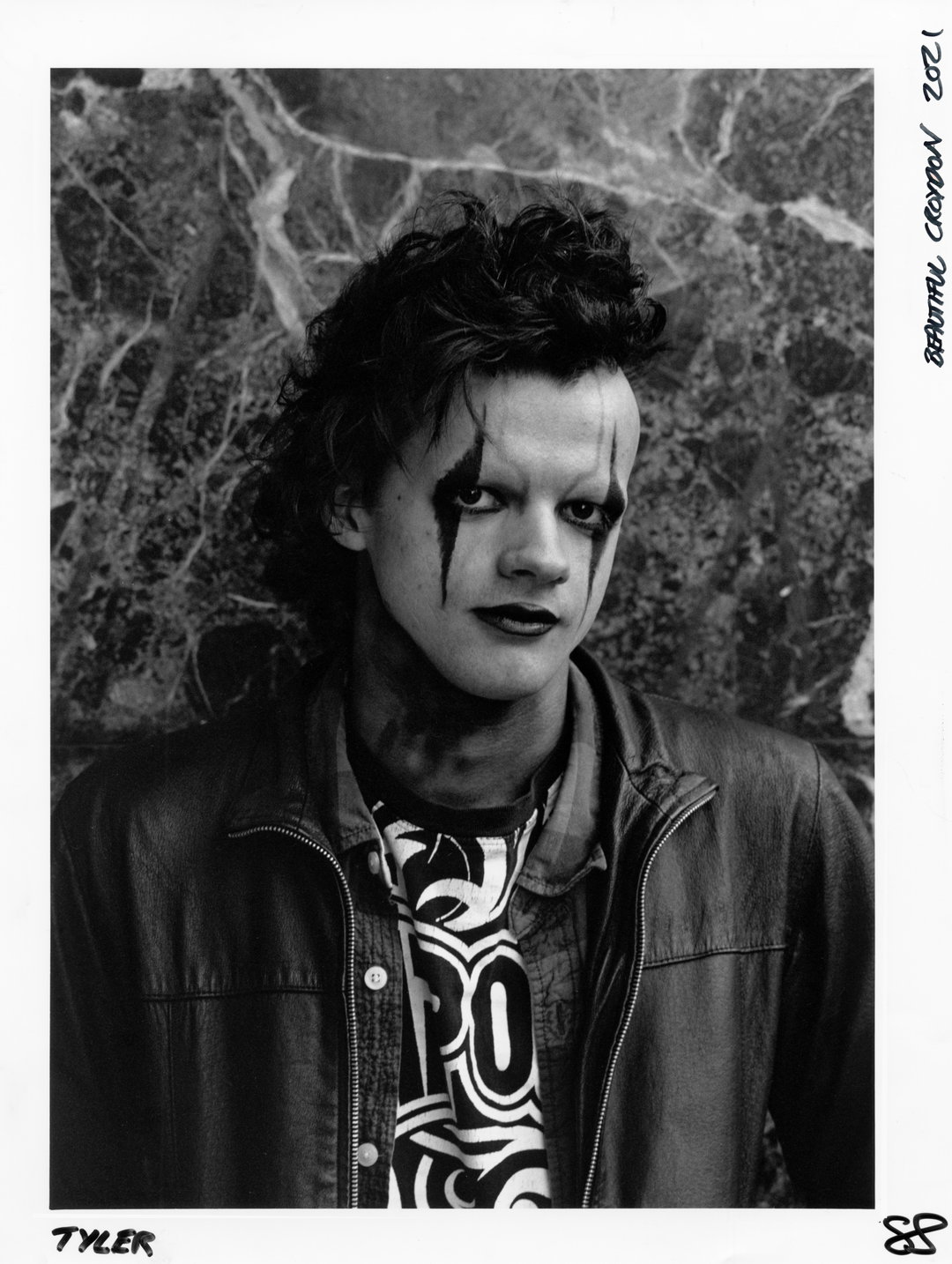 A black and white portrait of a person with dark, dramatic makeup around their eyes and lips. They wear a leather jacket over a T-shirt with a bold, swirling graphic.