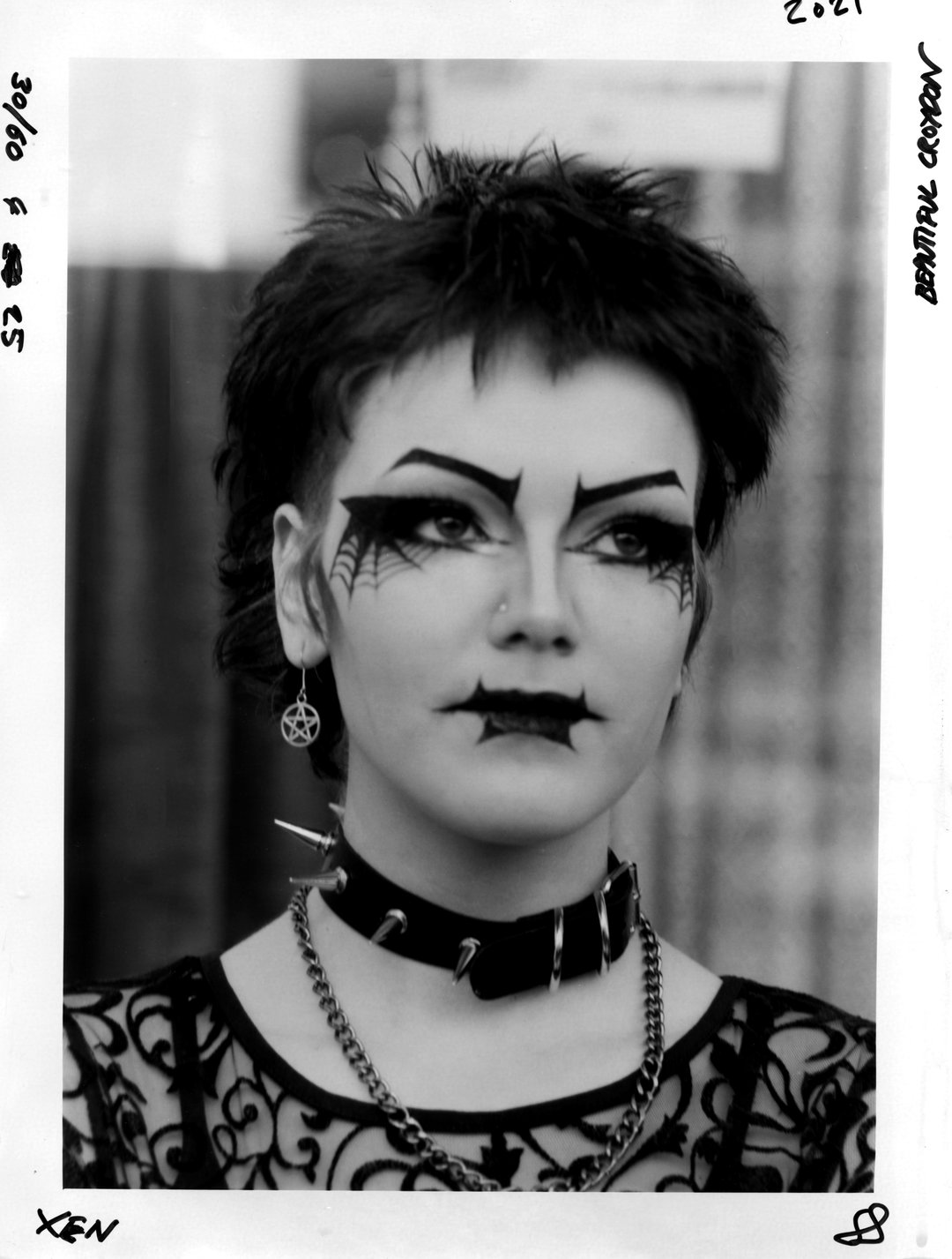 A person with short, spiky hair looks directly at the camera. They wear dramatic makeup including pointed eyebrows, spider-web patterns around the eyes, and a black lip design. A spiked leather collar and a silver chain are worn around their neck.
