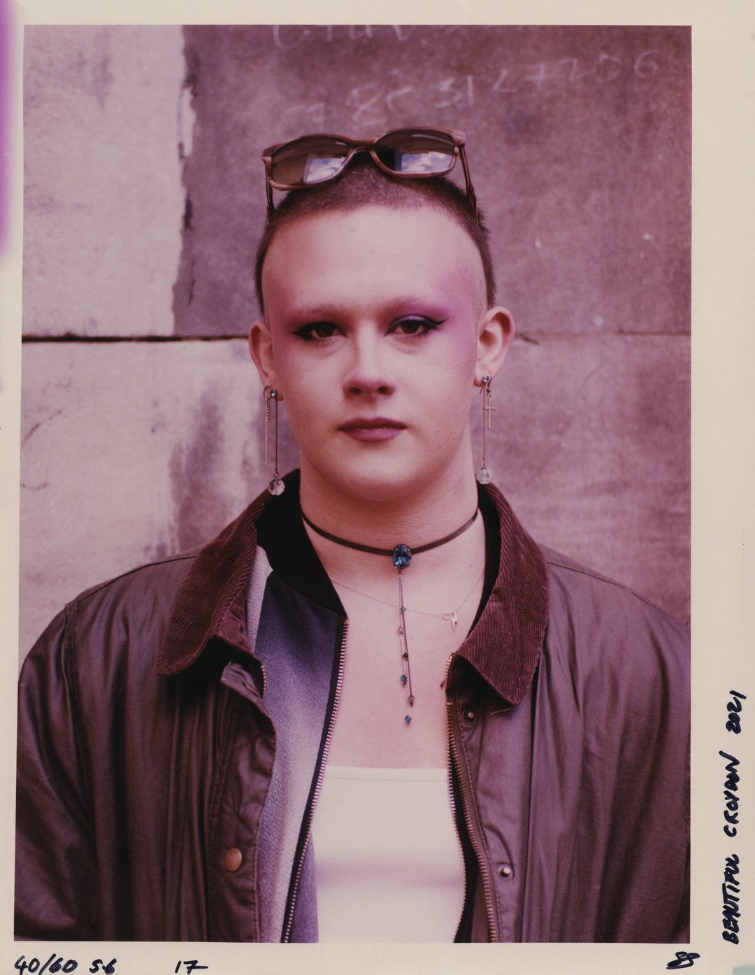 A person looks directly at the camera, wearing bold purple eyeshadow and long, dangling earrings. They wear a brown jacket over a white top, with sunglasses perched on their head.