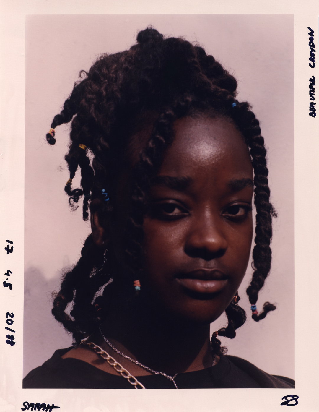 A person with twisted hair adorned with small colourful beads looks directly at the camera. The portrait is presented as a physical print with handwritten notes around the white border, including the name “Sarah”.