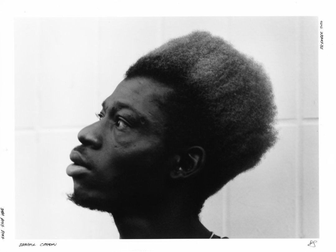 A black and white profile portrait of a person looking upwards. They have a high, rounded afro and a short beard.
