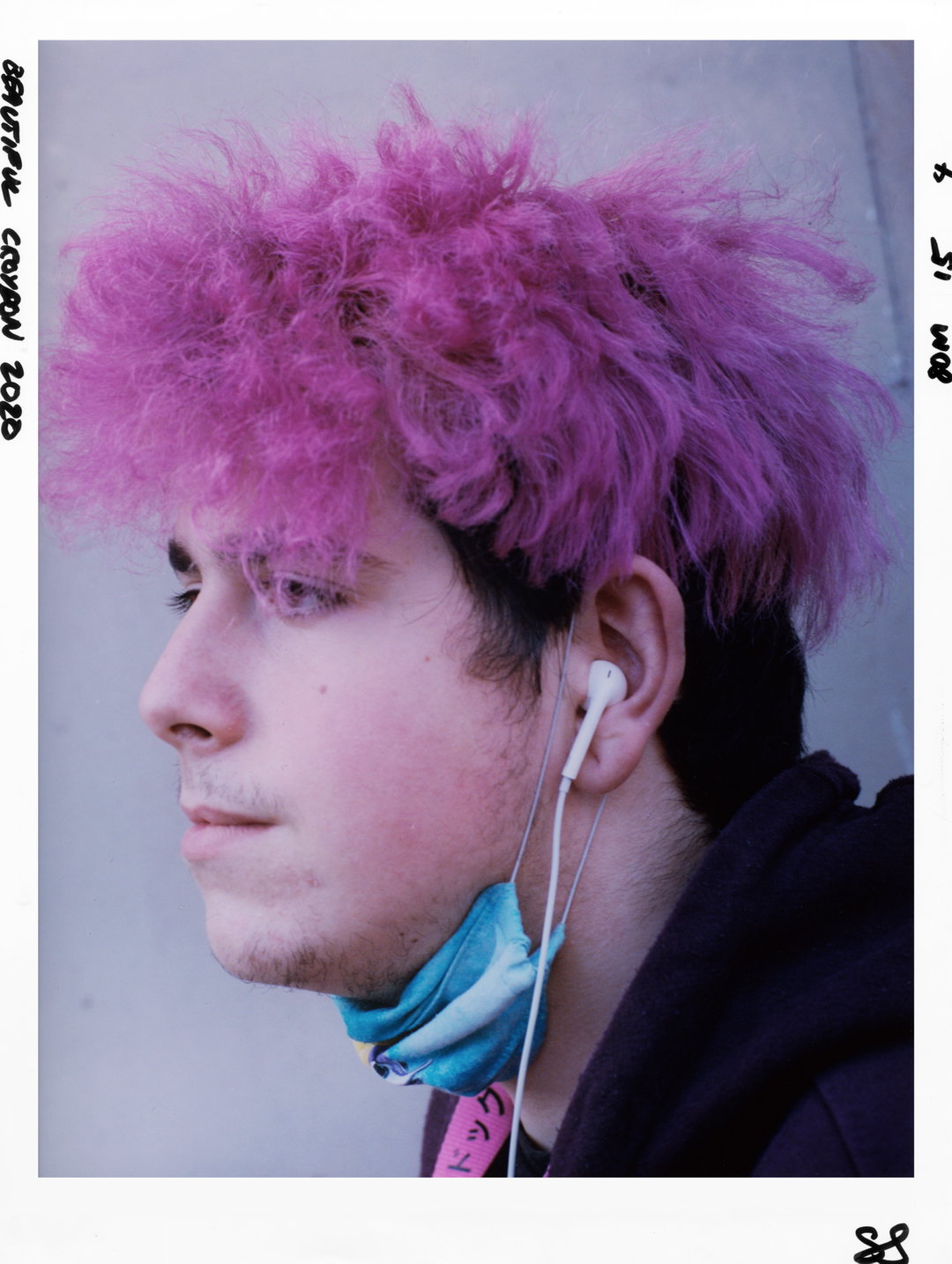 A person in profile with voluminous, bright pink hair and white earphones. A blue face mask is pulled down around their chin.