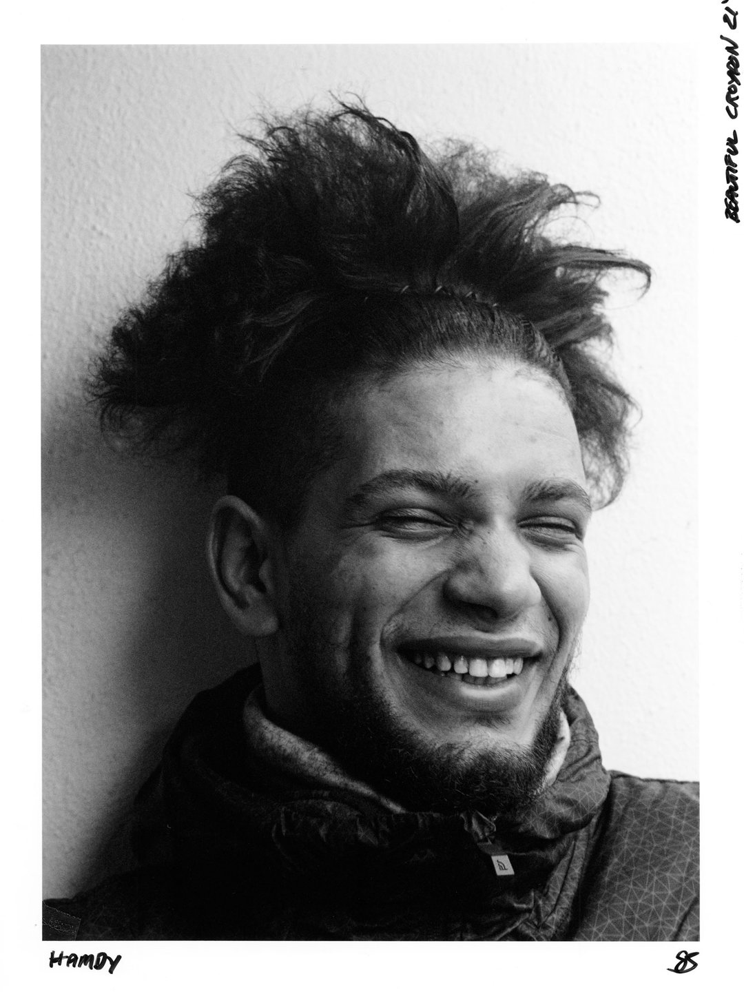 A black and white portrait of a person smiling with their eyes closed. They have voluminous, textured hair and a short beard, wearing a dark puffer jacket.