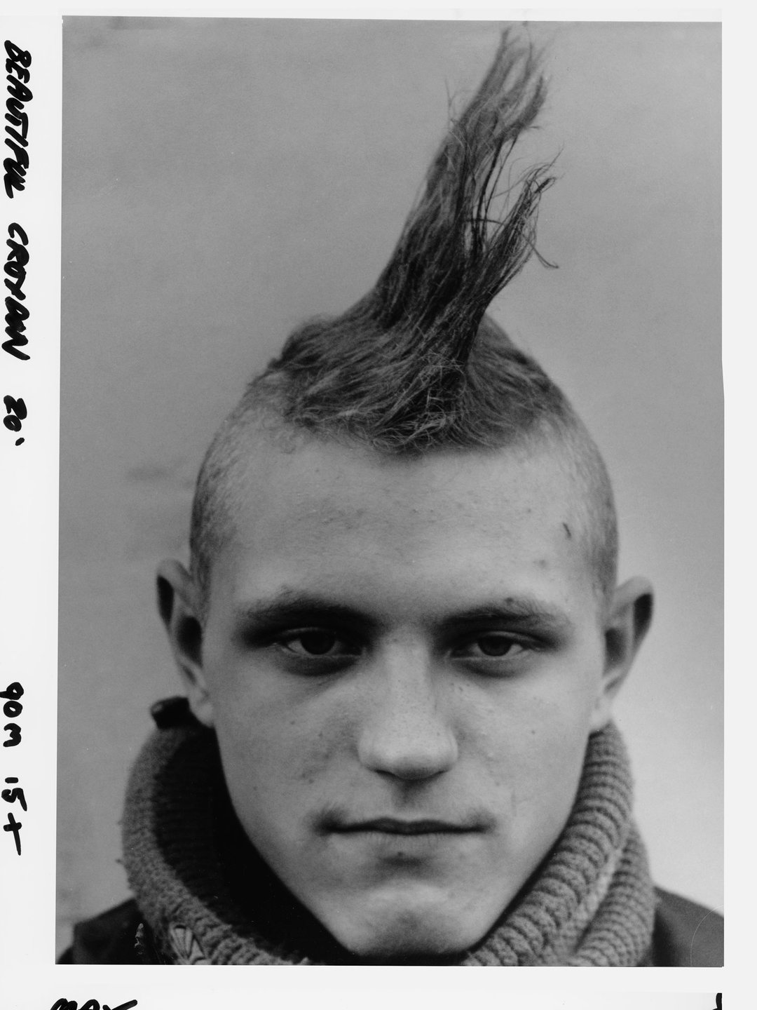 A black and white portrait of a person with a tall, thin mohawk. They wear a thick, ribbed knit collar and look directly at the camera.