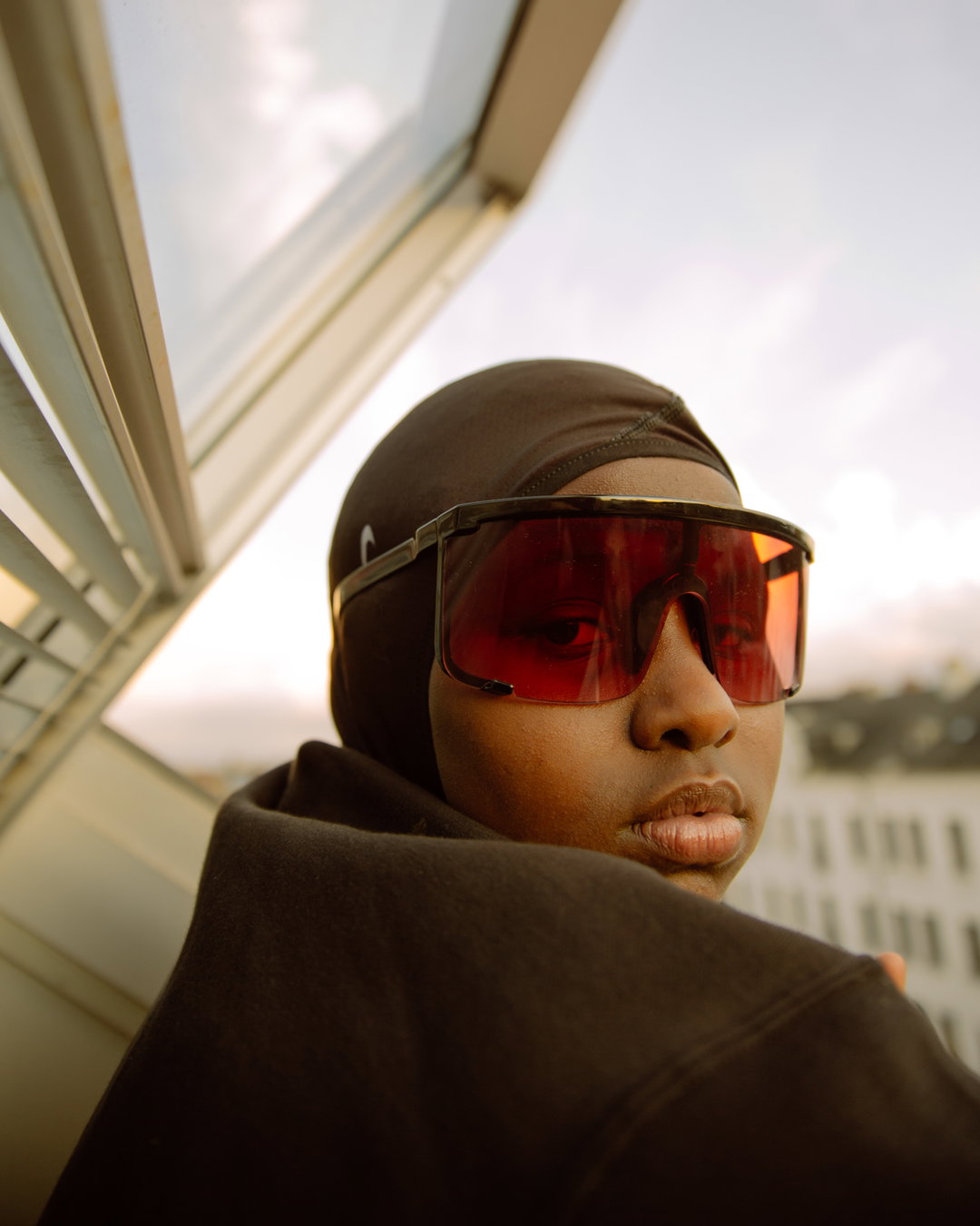 A person wearing a brown headscarf and large, red-tinted goggles looks over their shoulder. They are positioned against a pale sky and the edge of a white architectural structure.