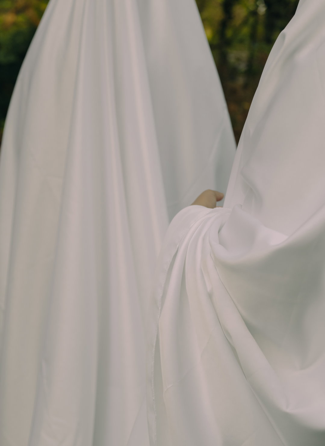 A close-up of two people draped in heavy, flowing white fabric. One person’s hand is visible, gently grasping the material of the other.