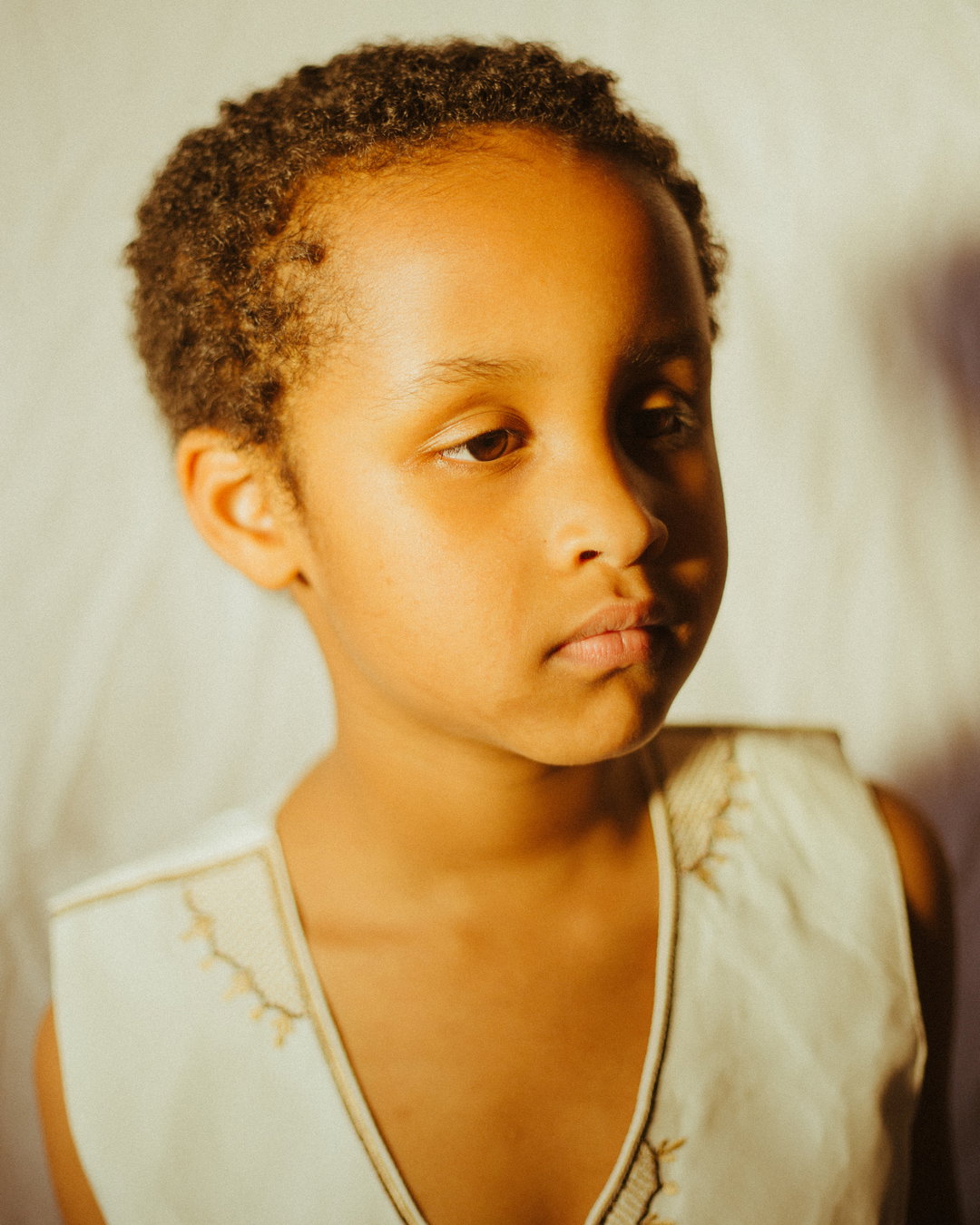 A child looks away from the camera with a neutral expression. They wear a white sleeveless top with gold embroidery against a plain, light background.