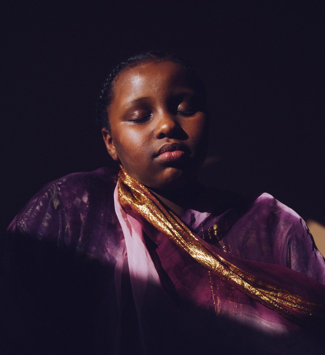 A person with their eyes closed, lit from the side against a dark background. They wear a purple garment with a shimmering gold trim.