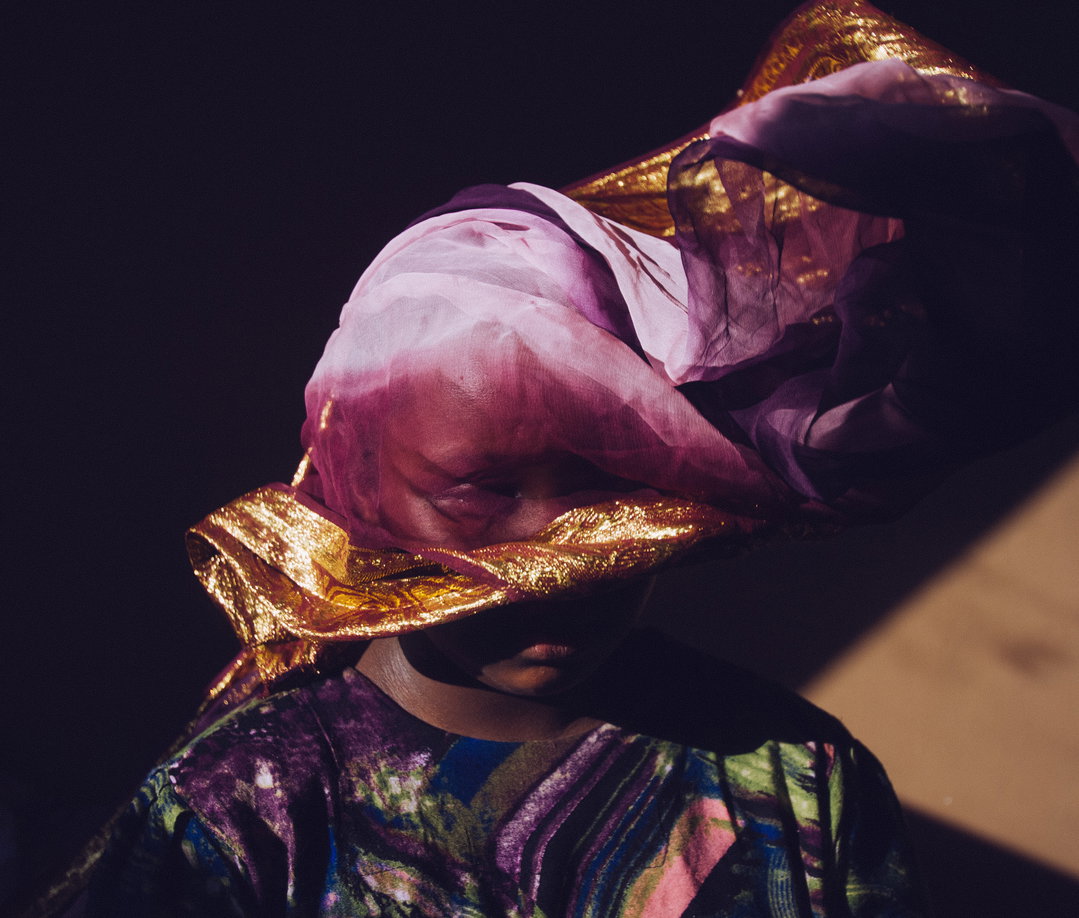 A person wears a voluminous headpiece made of flowing purple and gold fabric that obscures their face. They wear a matching iridescent garment with swirls of purple, blue and green.