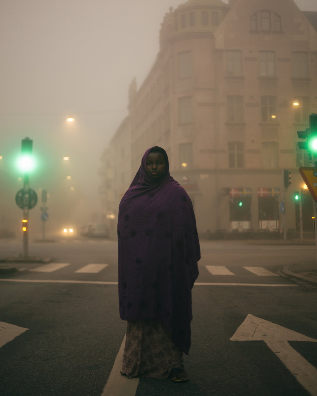 A person wearing a purple headscarf and wrap stands in the middle of a city road. Thick fog obscures the surrounding buildings and streetlights, with a green traffic light glowing to the left.