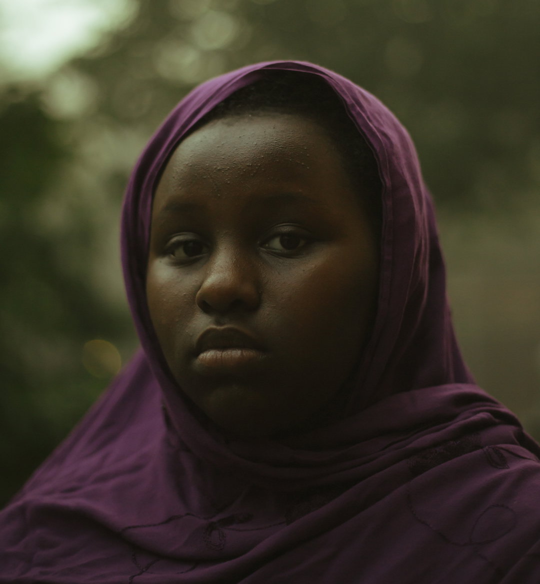 A person wearing a purple hijab looks directly at the camera with a neutral expression. The background is a soft, blurred green of foliage.