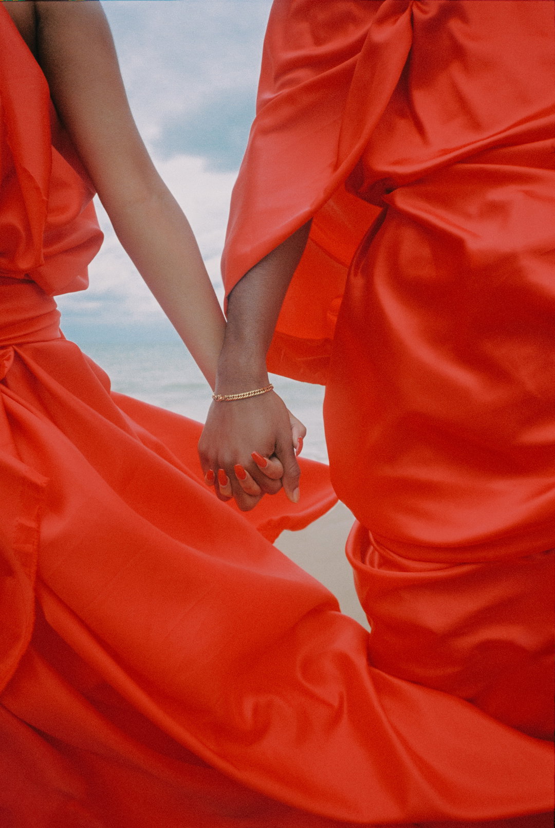 Two people hold hands, both wearing flowing, bright red satin garments. The fabric drapes and folds around them against a pale, cloudy sky.