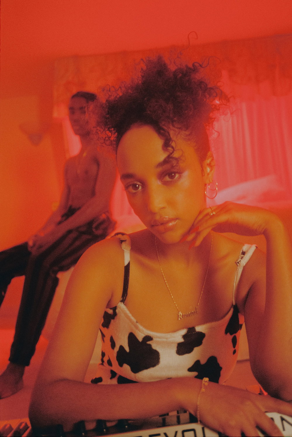 A person with curly hair and a cow-print top looks directly at the camera. Behind them, another person sits on a bed in a room bathed in deep red light.