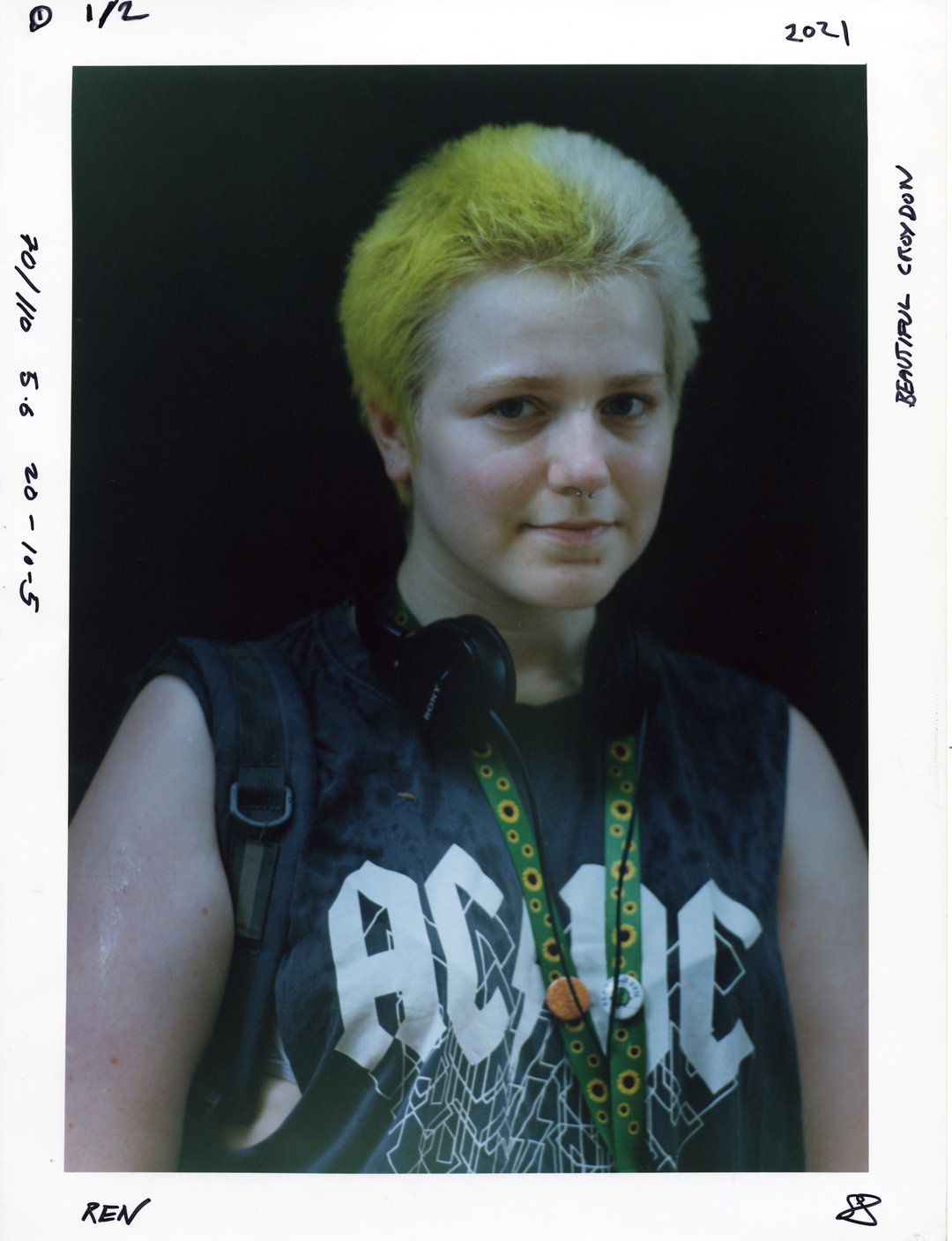 A person with short, yellow-dyed hair looks at the camera. They wear a black sleeveless top with the word “ACID” in bold white letters and have headphones around their neck.