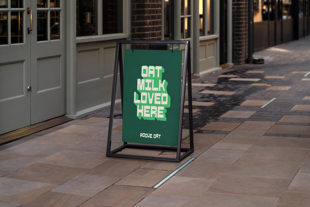 An A-frame sign on a paved pavement outside a shop. The green poster reads “OAT MILK LOVED HERE” in bold, 3D lettering, with “ROGUE OAT” written in smaller text below.