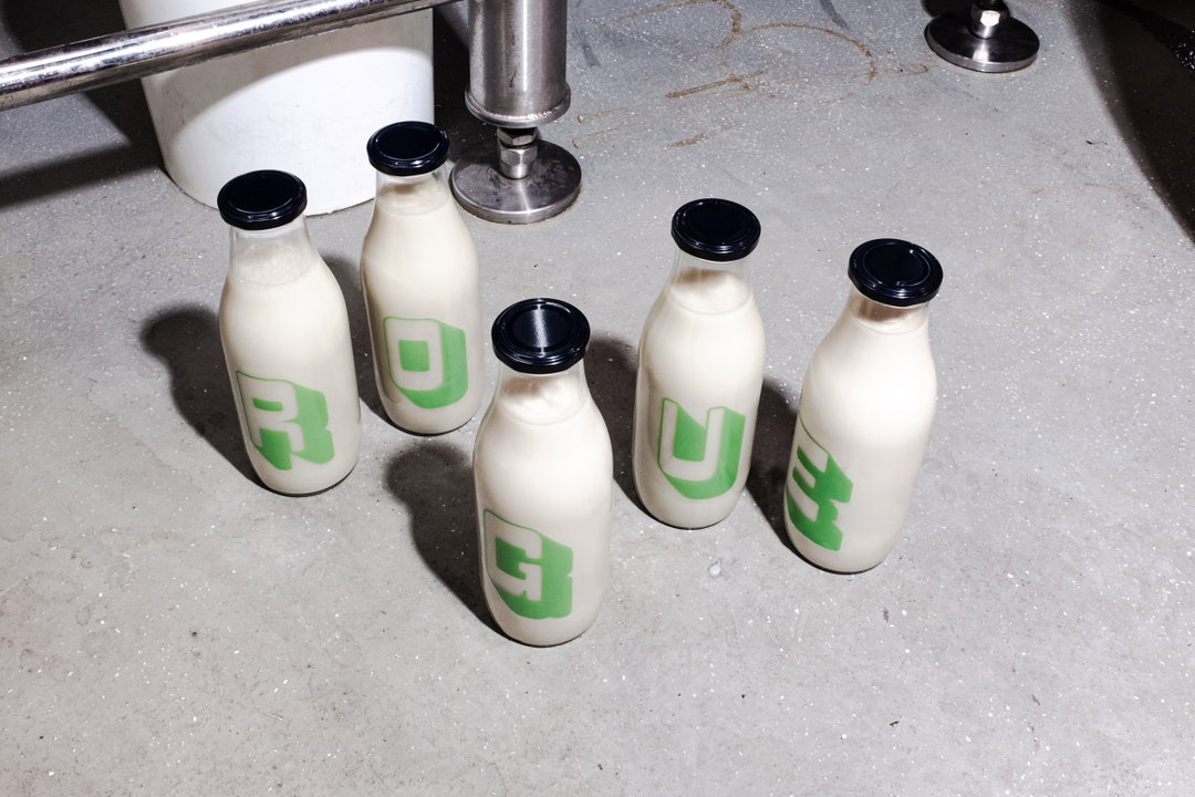 Five glass bottles filled with a white liquid stand on a grey concrete floor. Each bottle features a bold green letter, which together spell “Rogue”.