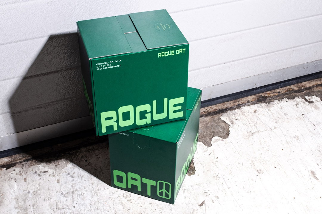 Two green cardboard boxes are stacked on a concrete floor against a white wall. The packaging features bold, lime green typography that reads “ROGUE” and “OAT”.
