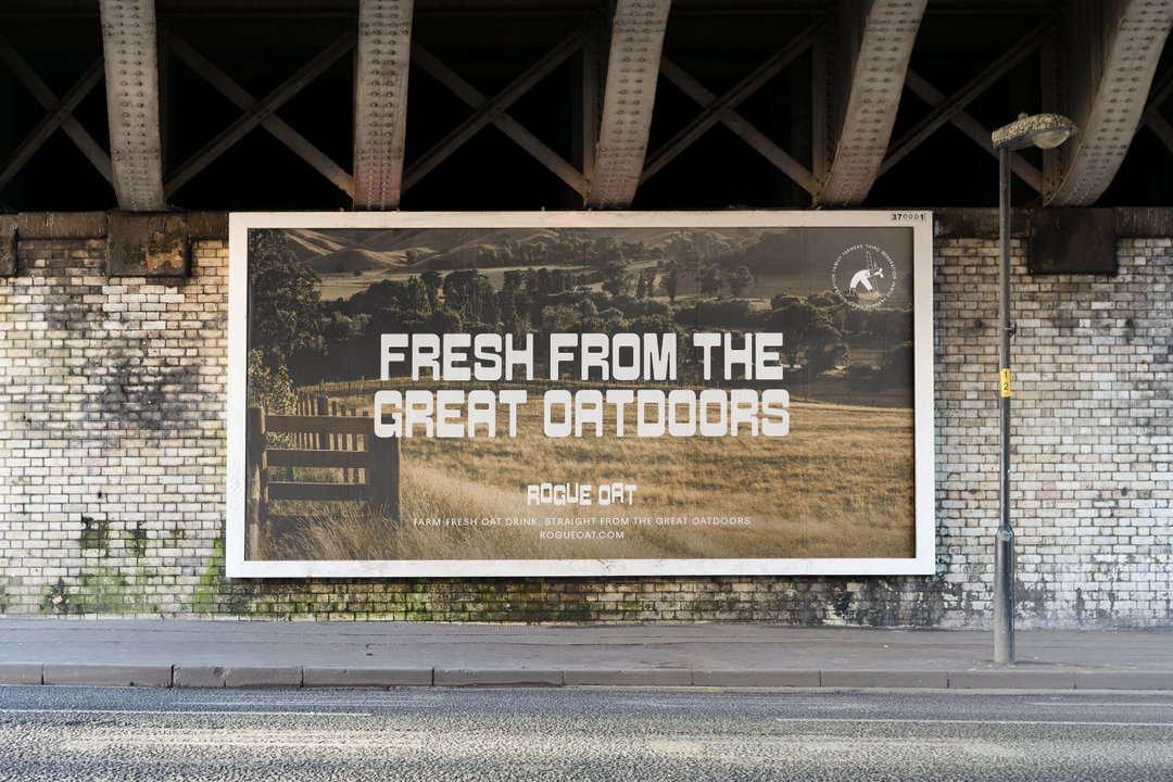 A large billboard is pasted onto a white brick wall beneath a railway bridge. The advertisement shows a rural landscape and reads “Fresh from the great outdoors” above the brand name “Rogue Oat”.