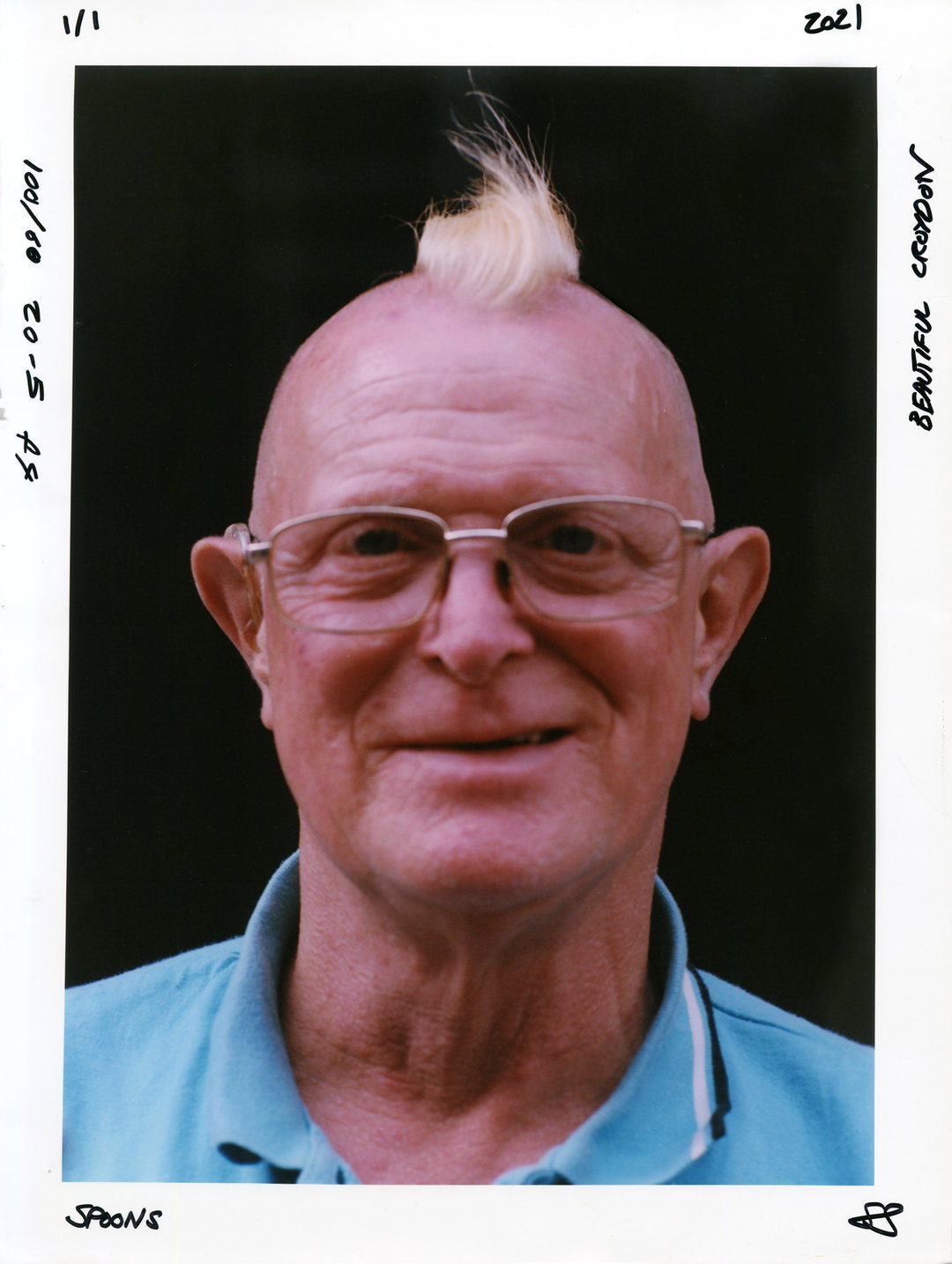 A person with glasses and a small blonde mohawk smiles at the camera. They wear a light blue polo shirt against a dark background. Handwritten notes on the white border include “Spoons” and “Beautiful Croydon”.