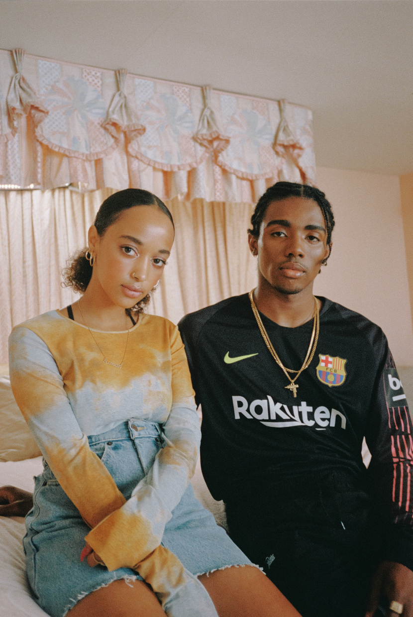 Two people sit close together on a bed in front of pale curtains. One wears a yellow and white tie-dye top with denim shorts, while the other wears a black FC Barcelona football shirt with “Rakuten” across the chest.
