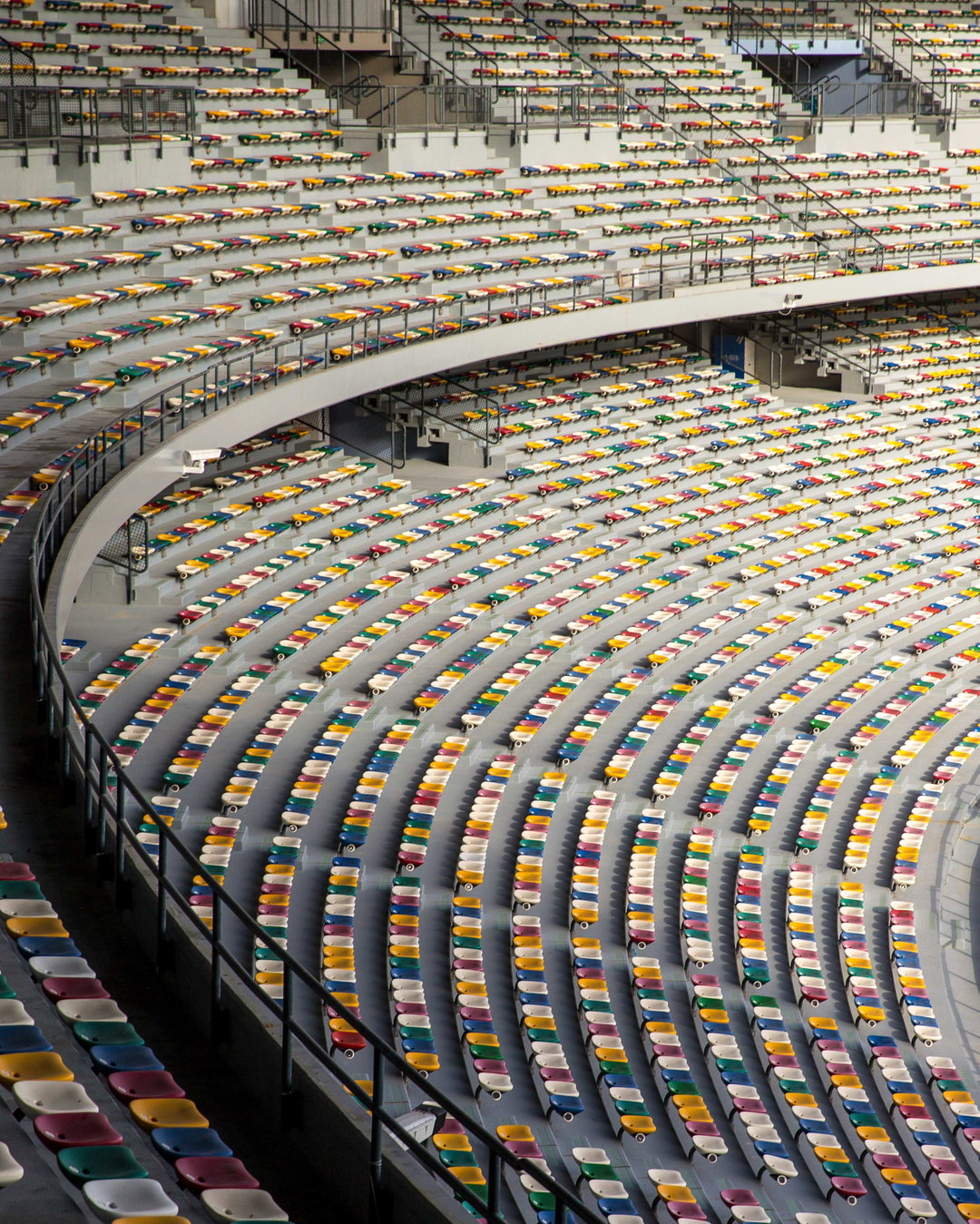 Rows of colourful stadium seats in yellow, red, green and blue curve across a concrete grandstand. The seats are arranged in sweeping arcs, creating a rhythmic pattern of bright colours against the grey architecture.