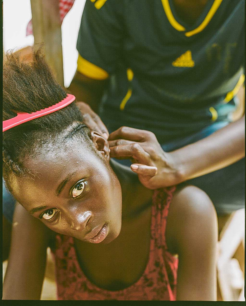 A person looks up at the camera while another person styles their hair. A pink comb is tucked into the hair, and the stylist's hands are positioned near the person's ear.
