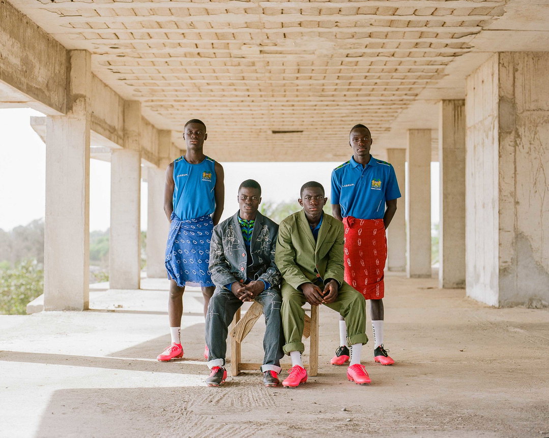 Four people pose together inside a concrete building shell with an open ceiling. They wear a mix of tailored jackets, blue shirts and colourful patterned wraps, paired with bright red shoes.