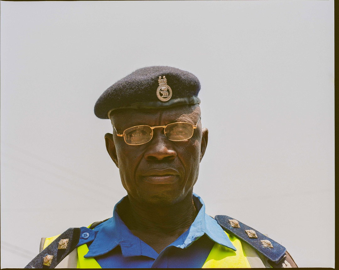 A person in a blue uniform and a black beret looks directly at the camera. They wear gold-rimmed glasses and a high-visibility yellow vest over their shoulders.