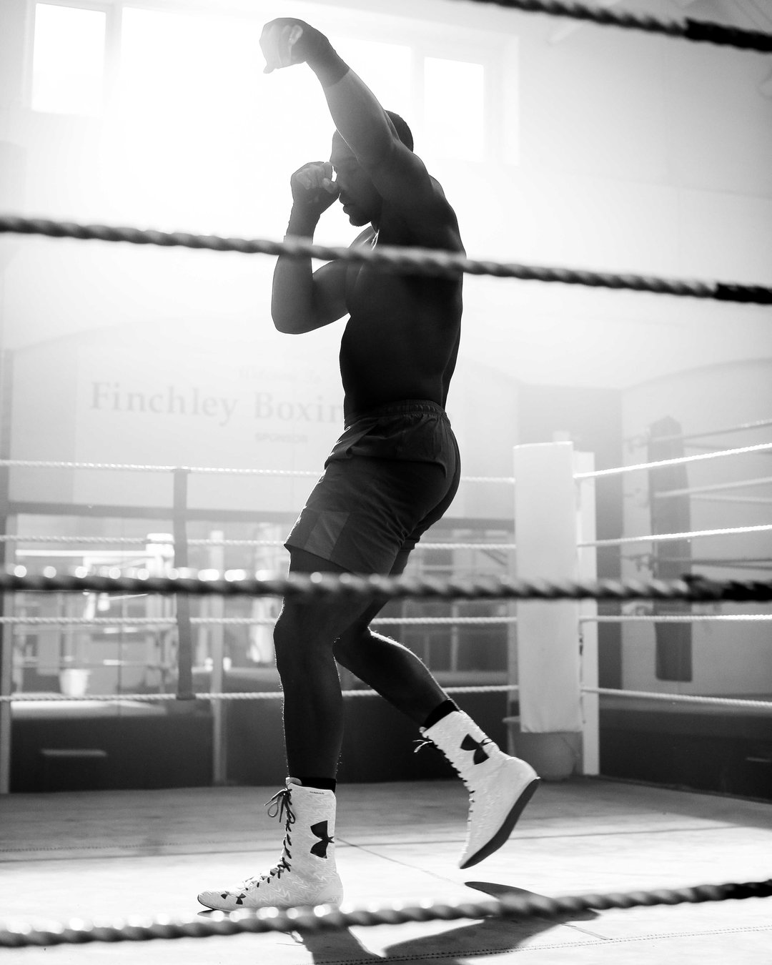 A black and white photo of a boxer in a boxing ring, captured in silhouette against a bright light. They stand in a fighting stance, wearing boxing boots and shorts, with the words “Finchley Boxing” visible on the wall behind them.