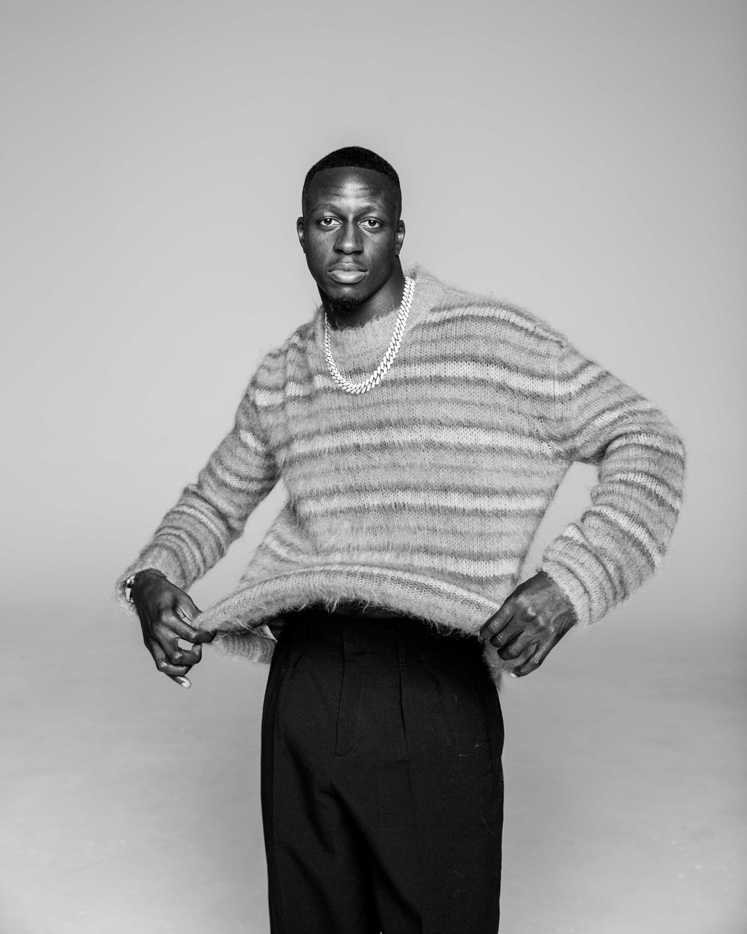 A person stands against a plain background, looking directly at the camera. They wear a fuzzy, striped knit jumper, dark trousers and a chunky chain necklace.
