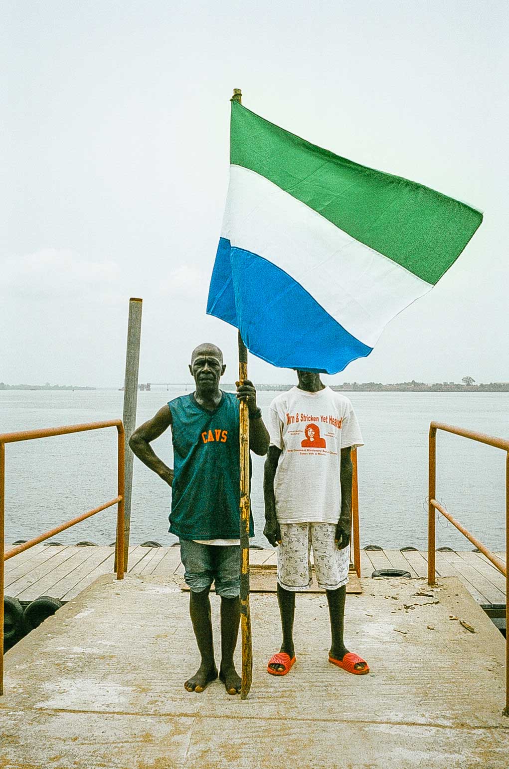 Two people stand on a concrete pier, holding a large green, white and blue flag between them. One person wears a dark green “Cavs” vest and denim shorts, while the other wears a white T-shirt and patterned shorts.