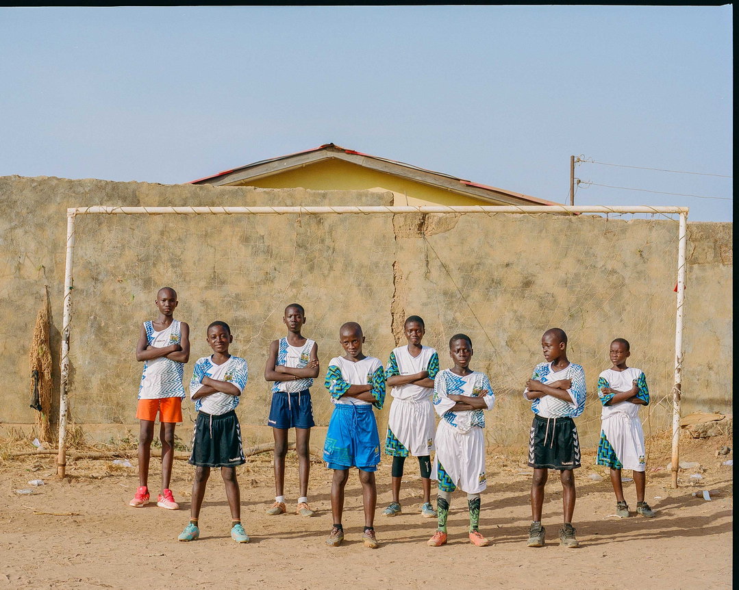Nine children stand in a line in front of a football goal on a dusty pitch. They wear matching blue and white sports kits and pose with their arms crossed.