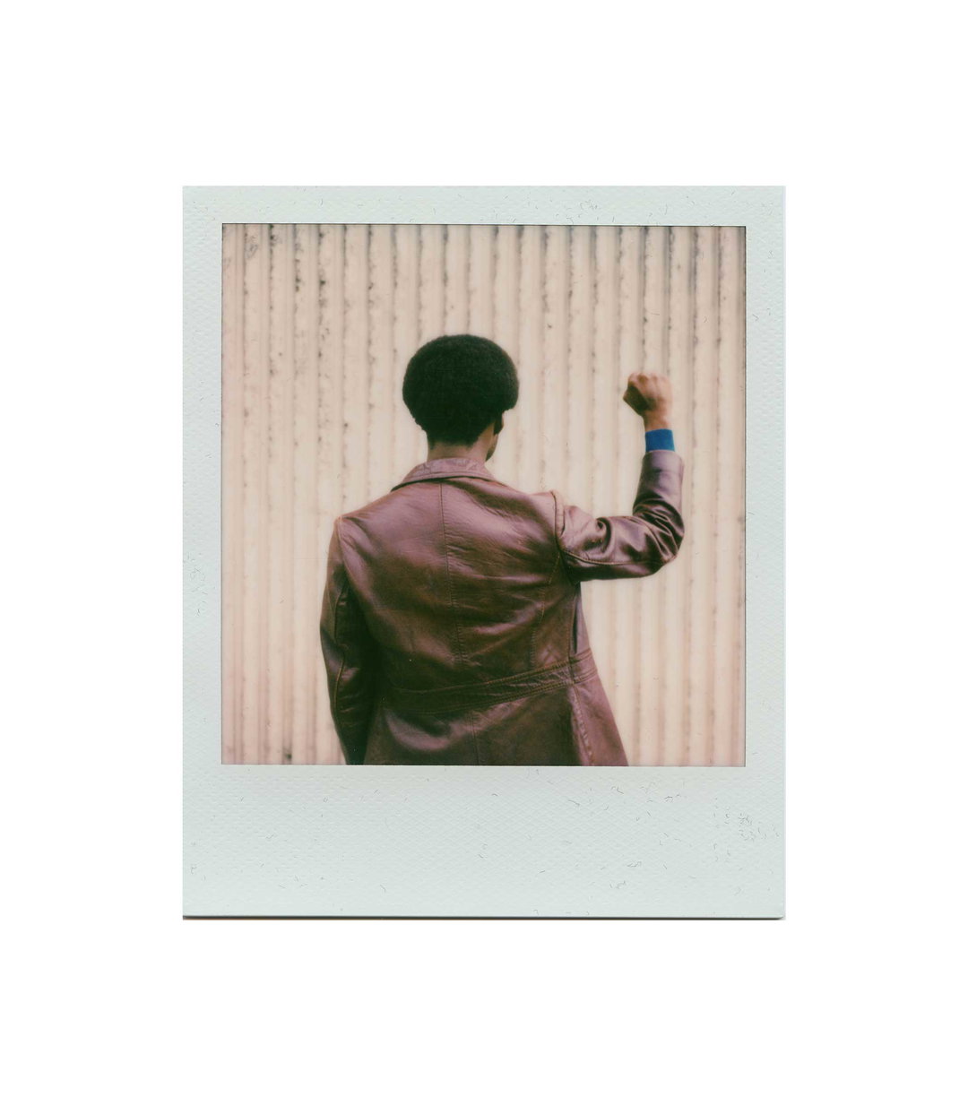 A person with an afro is seen from behind, wearing a brown leather jacket. They have one arm raised with a clenched fist against a backdrop of a corrugated wall.
