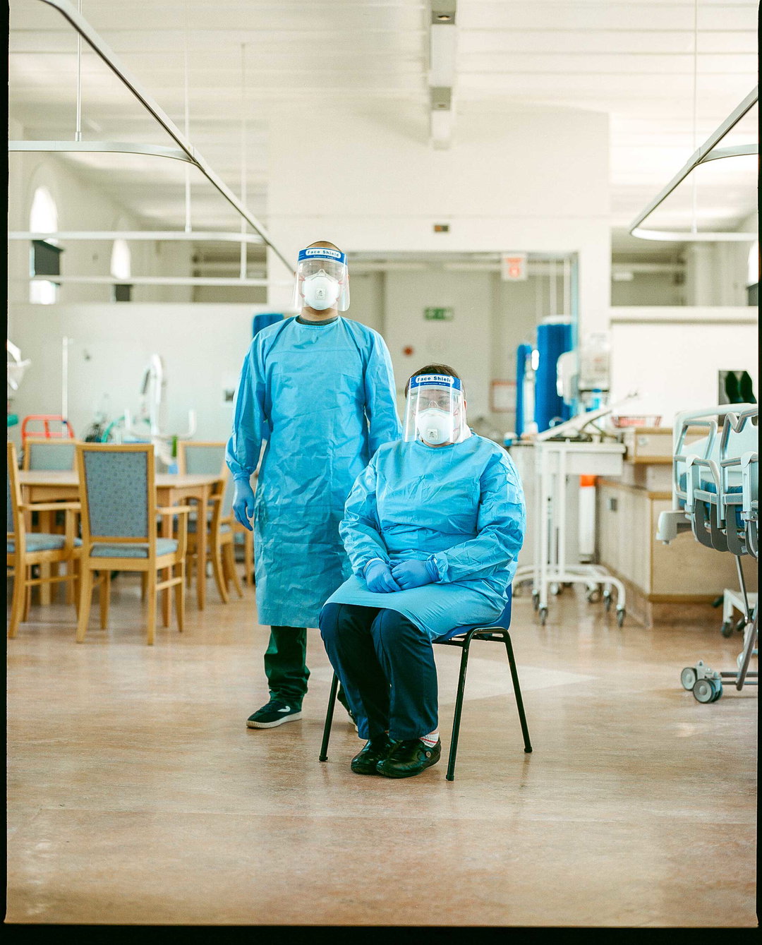 Two people in blue medical gowns, face shields and masks stand in a bright hospital ward. One person stands while the other sits on a chair, both looking towards the camera.