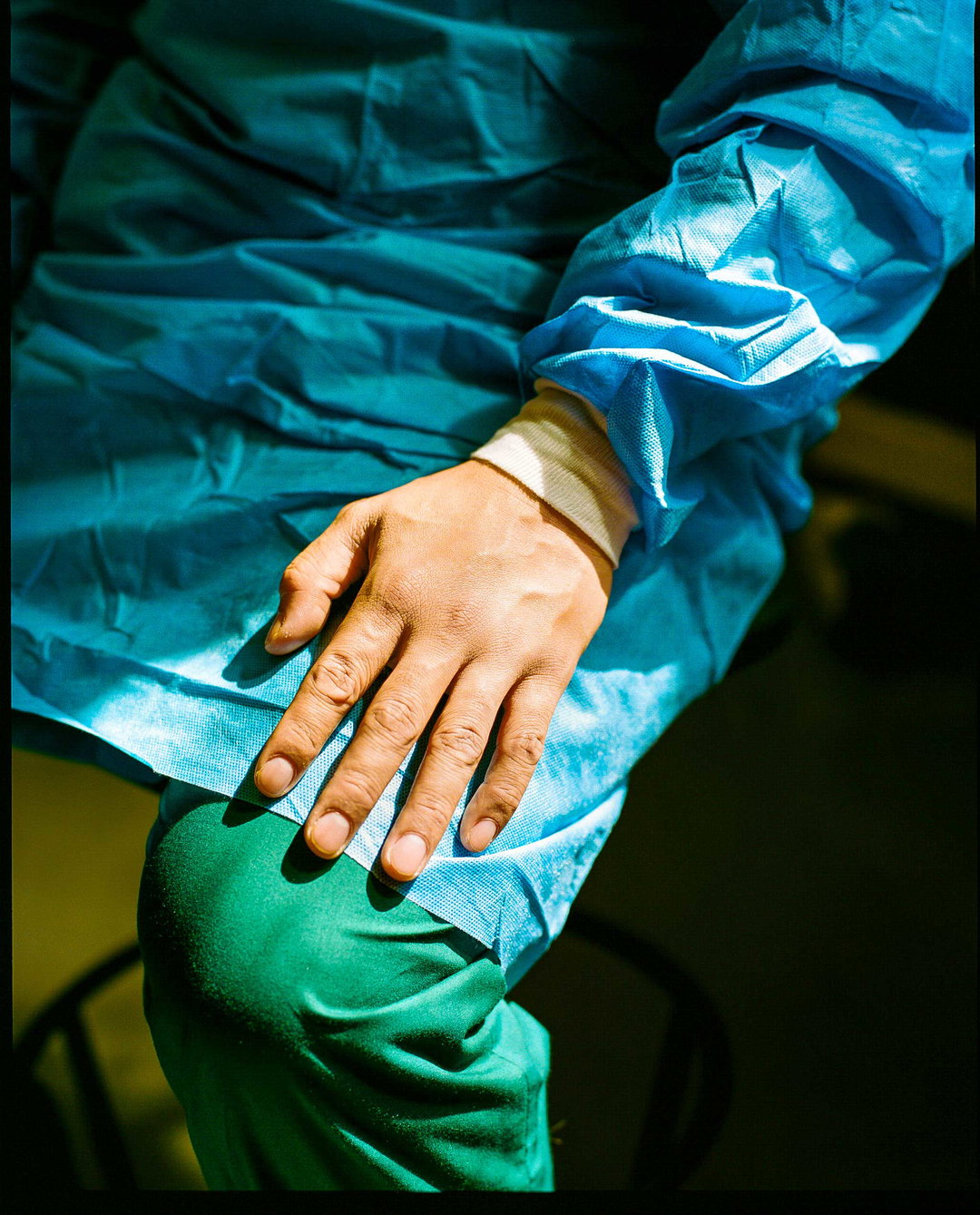 A close-up of a person wearing blue surgical scrubs and green trousers. Their hand rests on their lap, lit by a sharp beam of light.