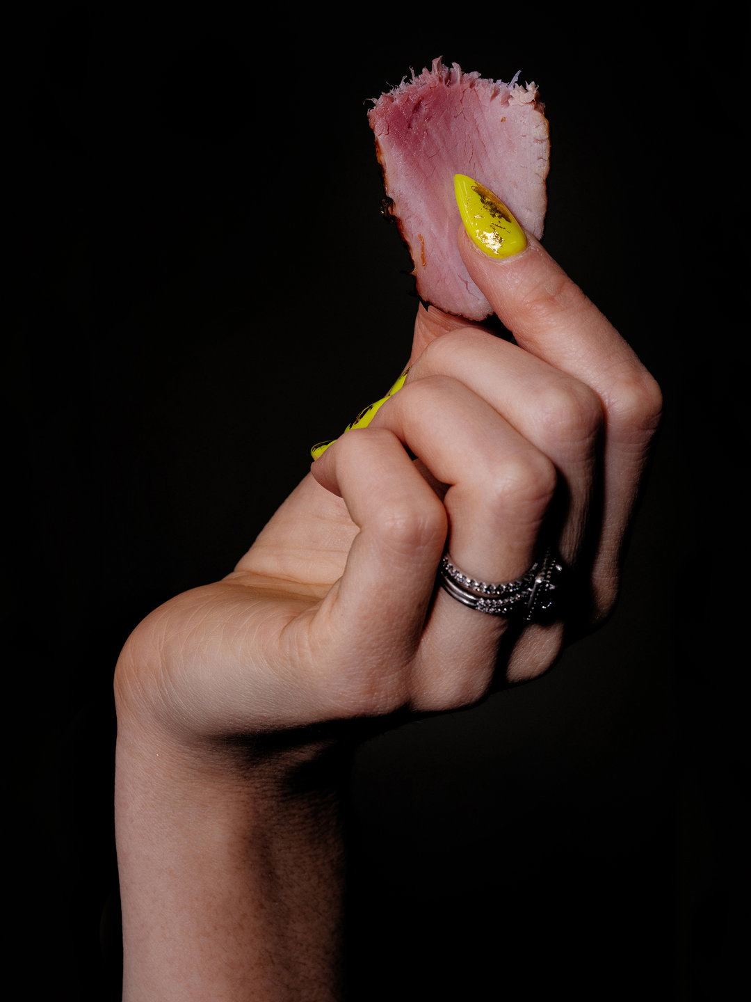 A hand with long, bright yellow nails holds a thin slice of pink meat against a black background. Several silver rings are worn on the ring finger.