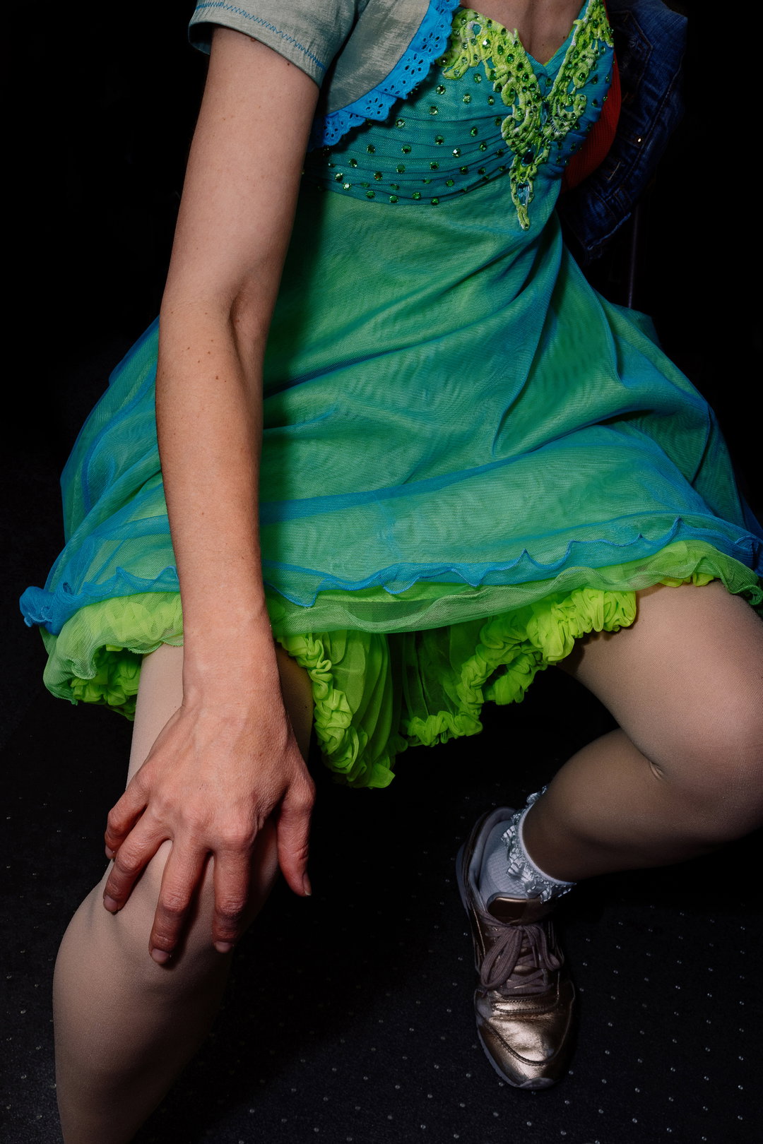 A person crouches against a dark background, wearing a layered green and blue dress with a ruffled hem. They wear white tights and metallic gold trainers.