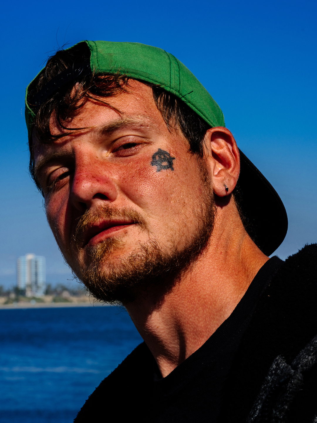 A person wearing a green baseball cap looks directly at the camera. They have a small star tattoo on their left cheek and a short beard.