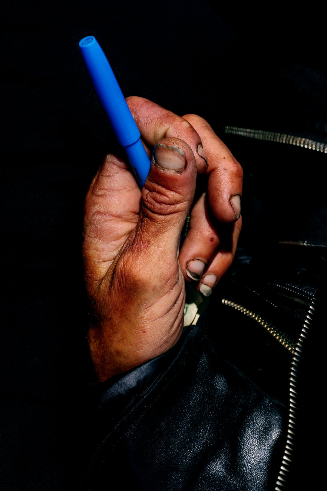 A close-up of a hand holding a bright blue pen against a dark background. The person wears a black leather jacket with a silver zip.