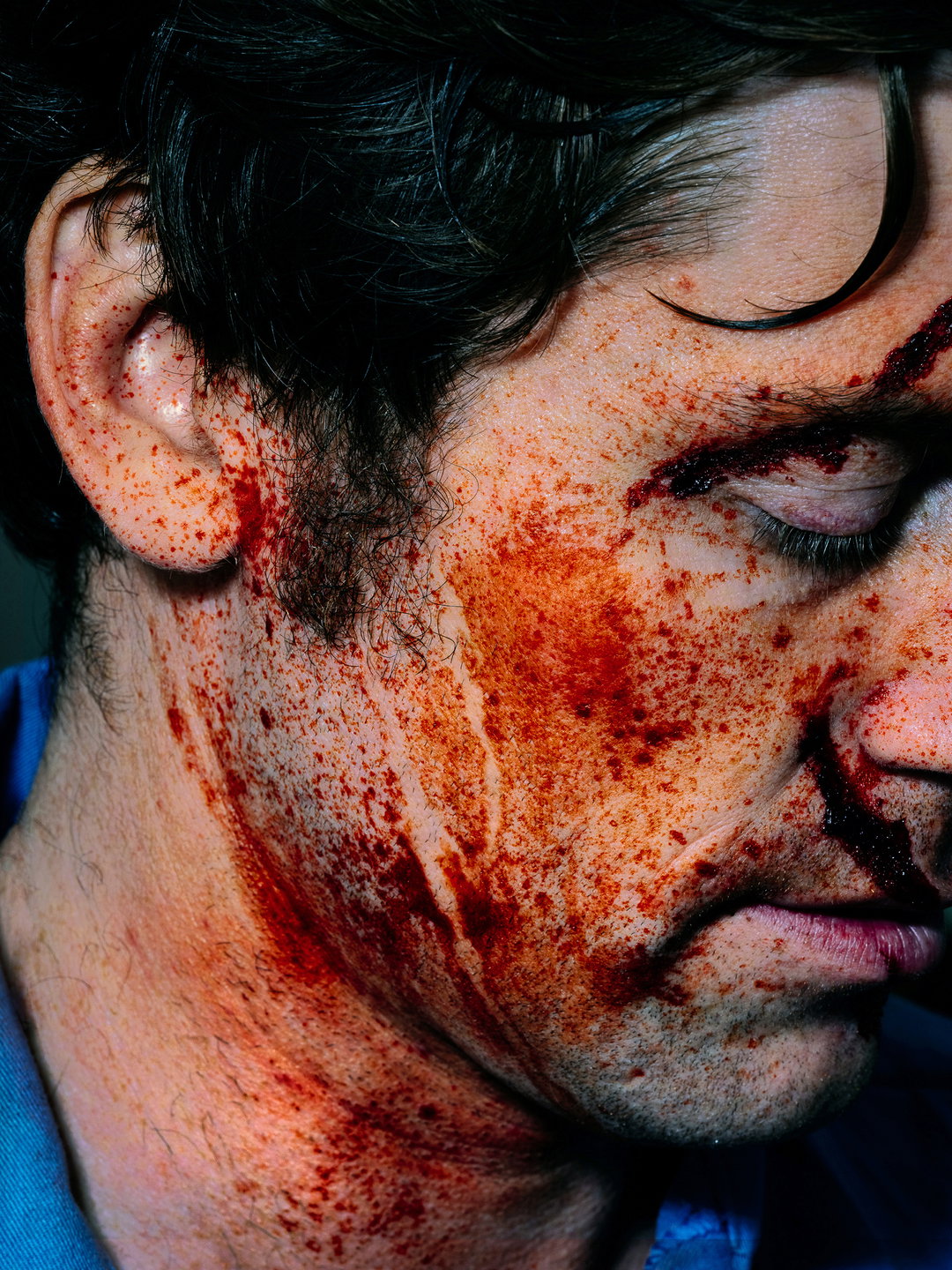 A close-up of a person's face covered in blood and grime. They have their eyes closed and a deep cut across their right eyelid.
