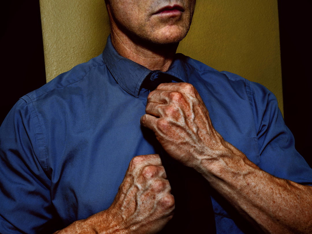 A person in a blue shirt tightens a dark tie. Their hands are clenched, with prominent veins visible across their knuckles and wrists.